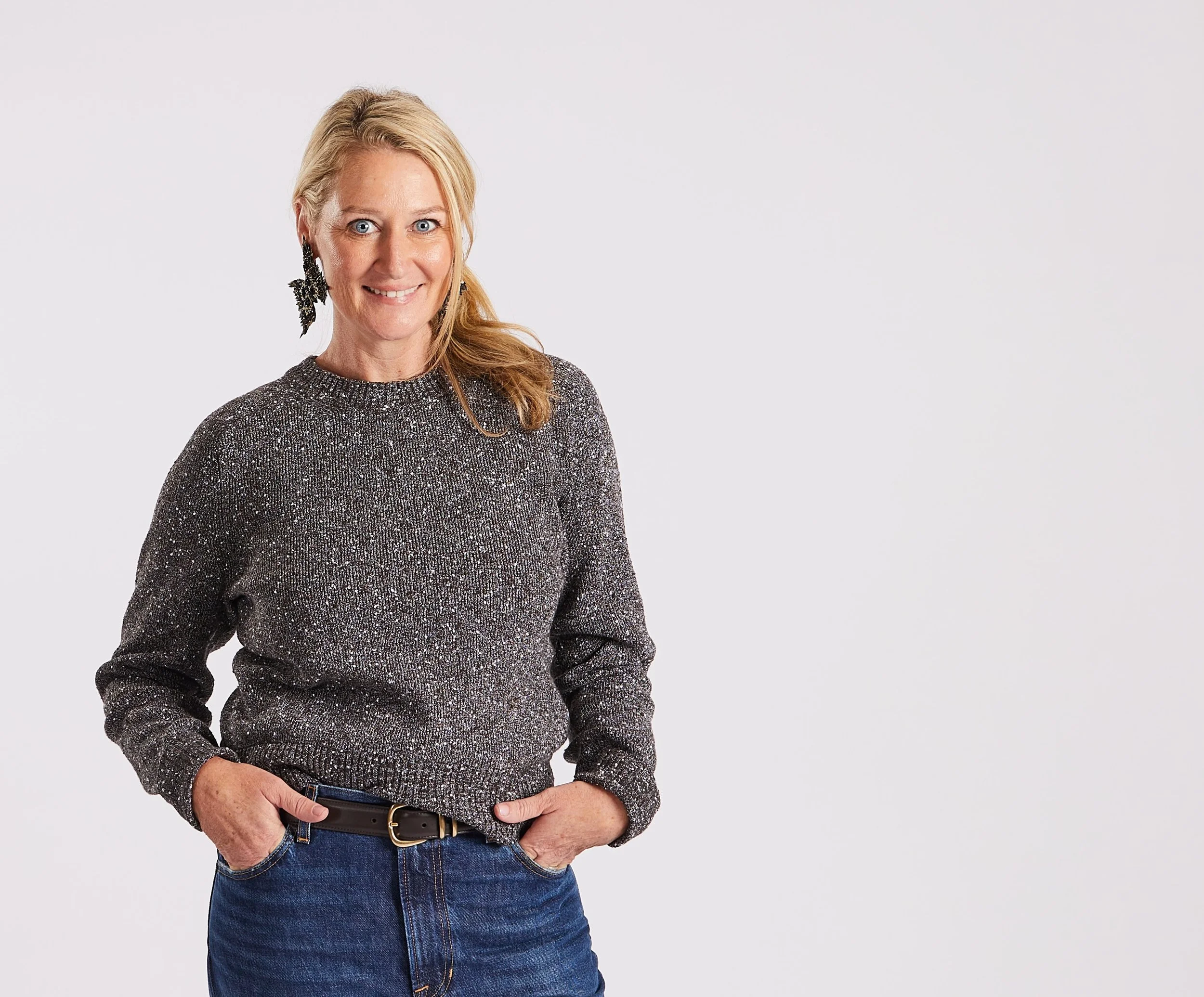 A woman wearing a sparkly gray sweater, blue jeans, and a black belt, with blonde hair, standing against a plain white background.