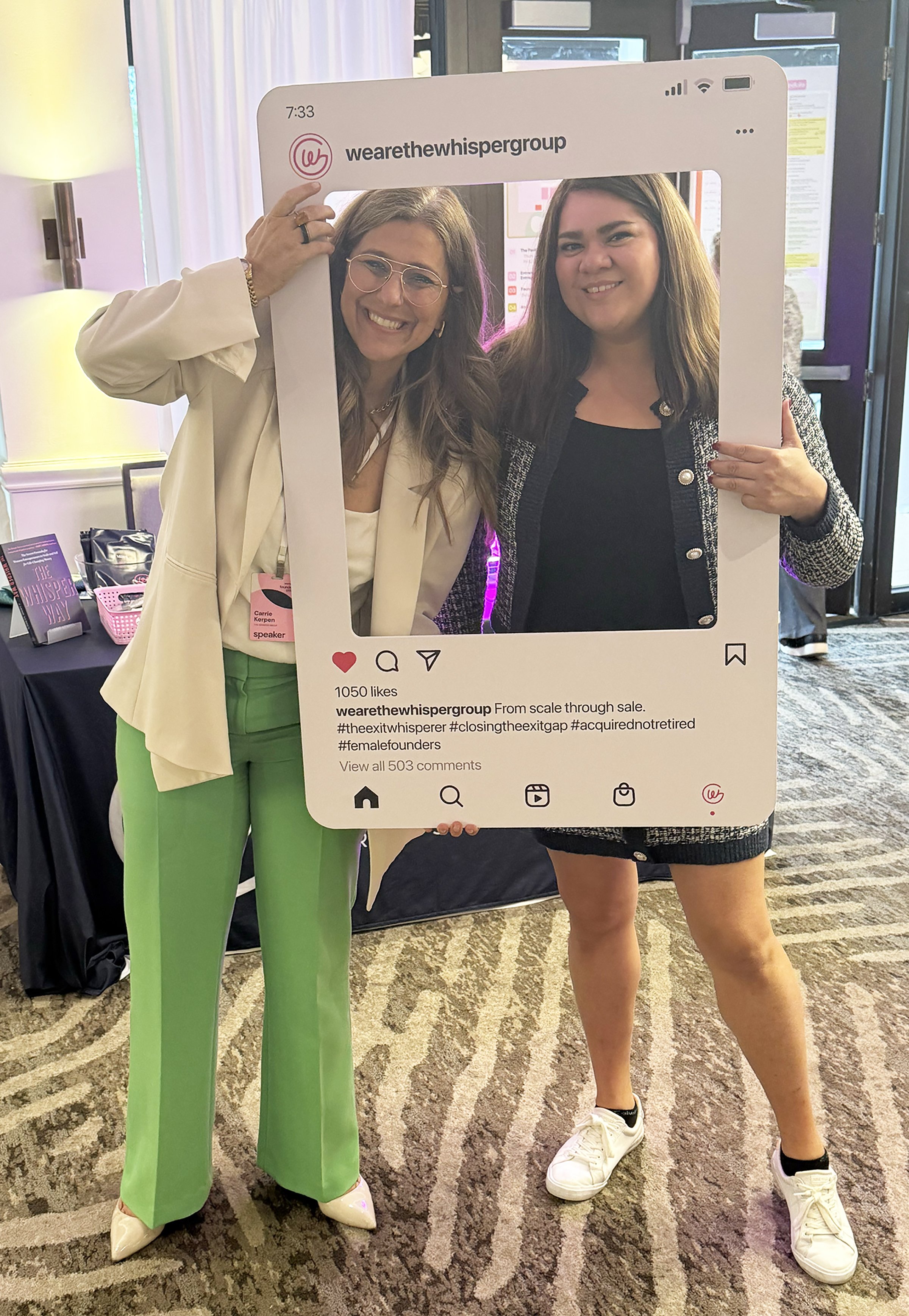 Two women holding a large social media frame prop at a conference. The woman on the left wears a cream blazer, green pants, and beige shoes, with glasses and a name tag. The woman on the right wears a black dress with a patterned jacket and white sneakers. The background shows conference materials and a patterned carpet.