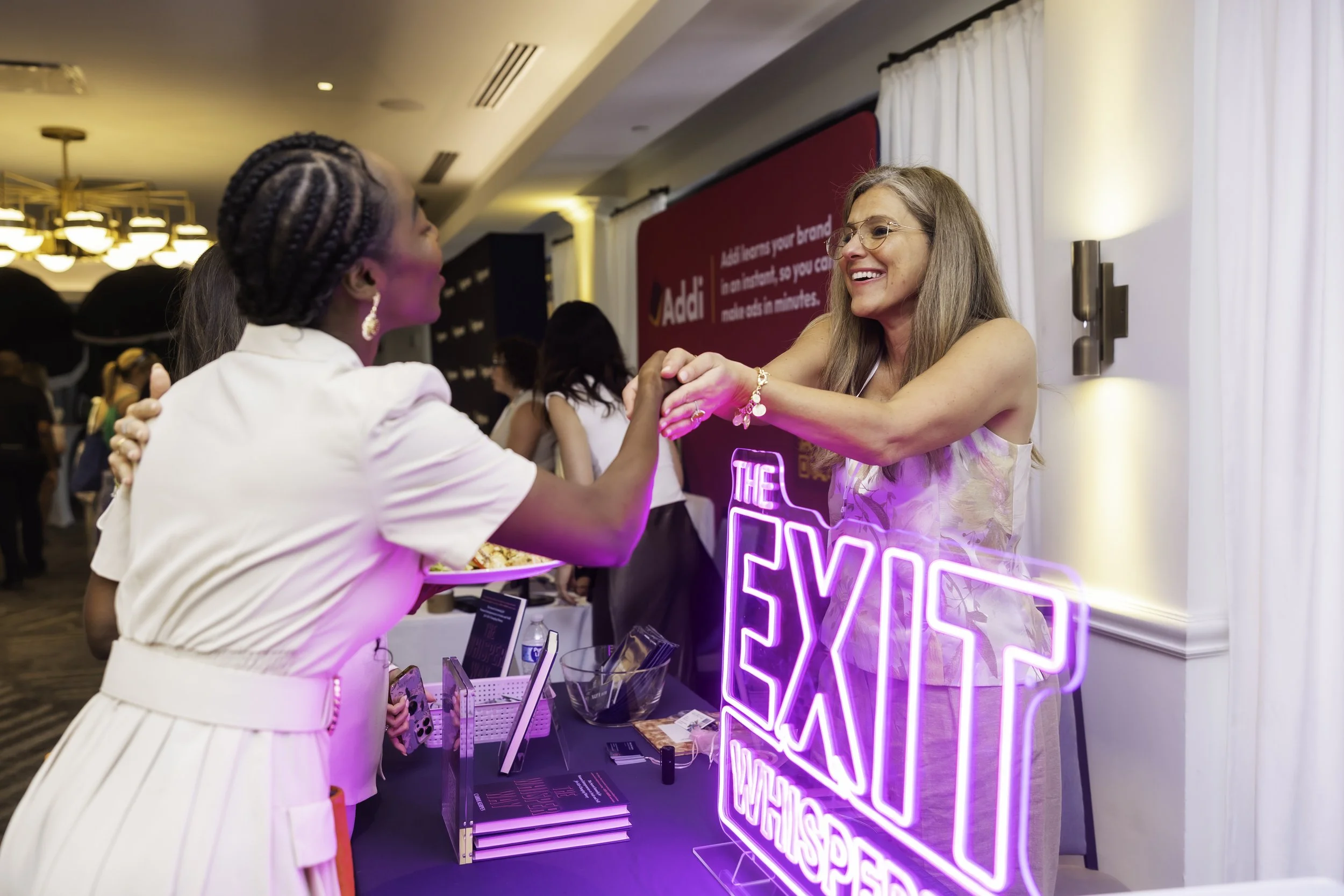 A woman with glasses and grey hair talking to a woman with braided hair at a booth with a neon sign that says 'LOVE' and a pink background in what appears to be a conference or event space.
