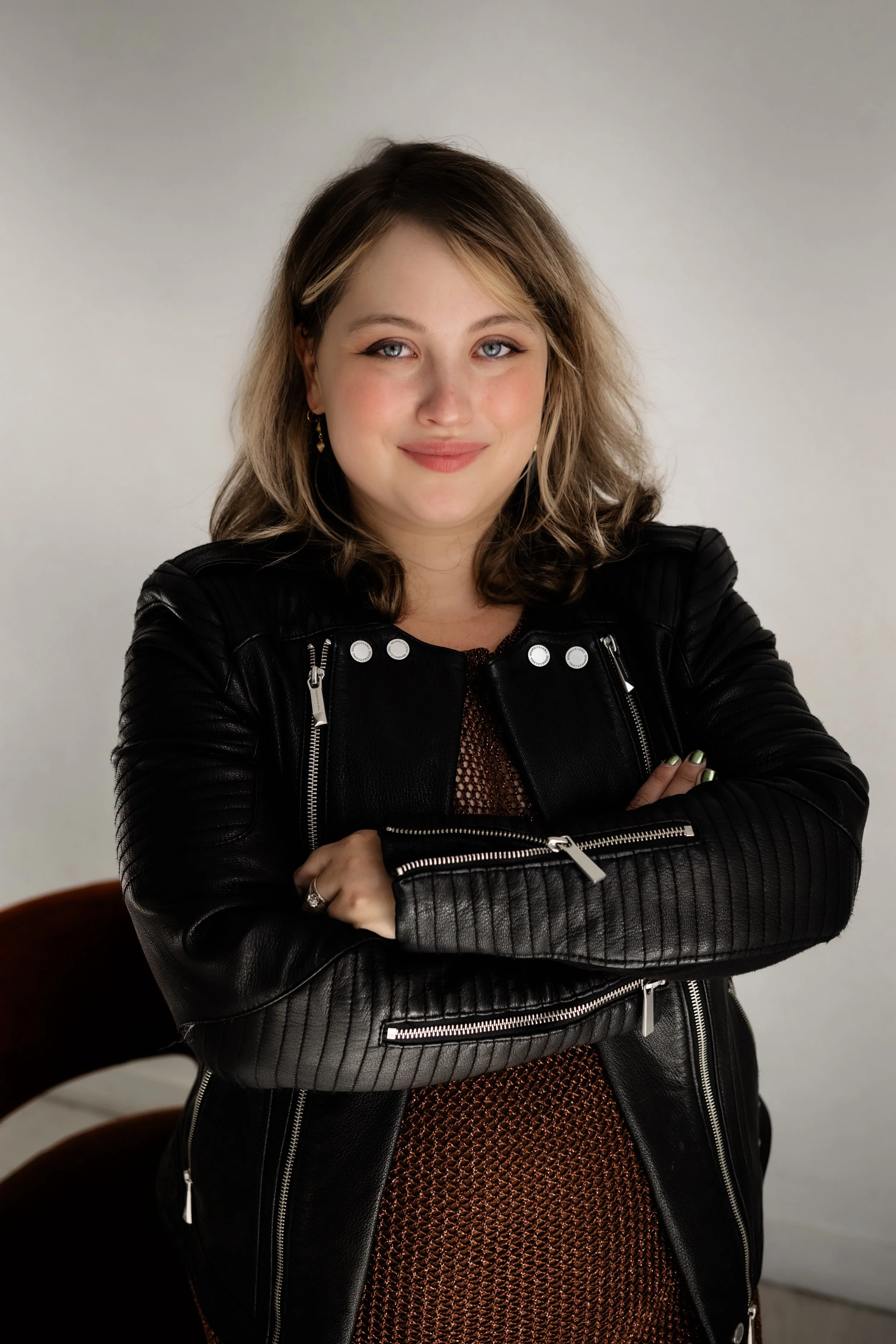 A woman with light brown hair and blue eyes, smiling with arms crossed, wearing a black leather jacket and a brown patterned dress, sitting in front of a plain light-colored wall.