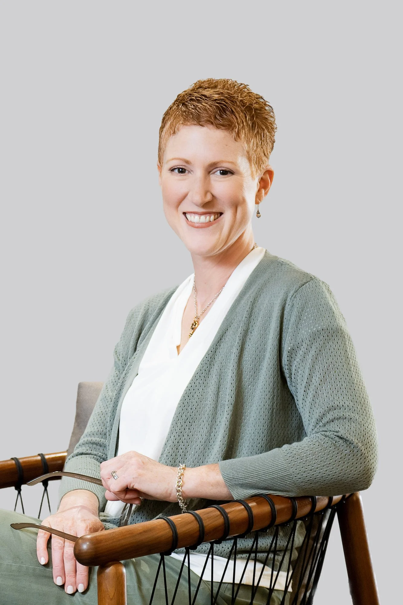 A woman with short red hair smiling while sitting on a wooden chair with a black wire frame, wearing a gray cardigan over a white blouse, with her hands resting on her lap and holding sunglasses.