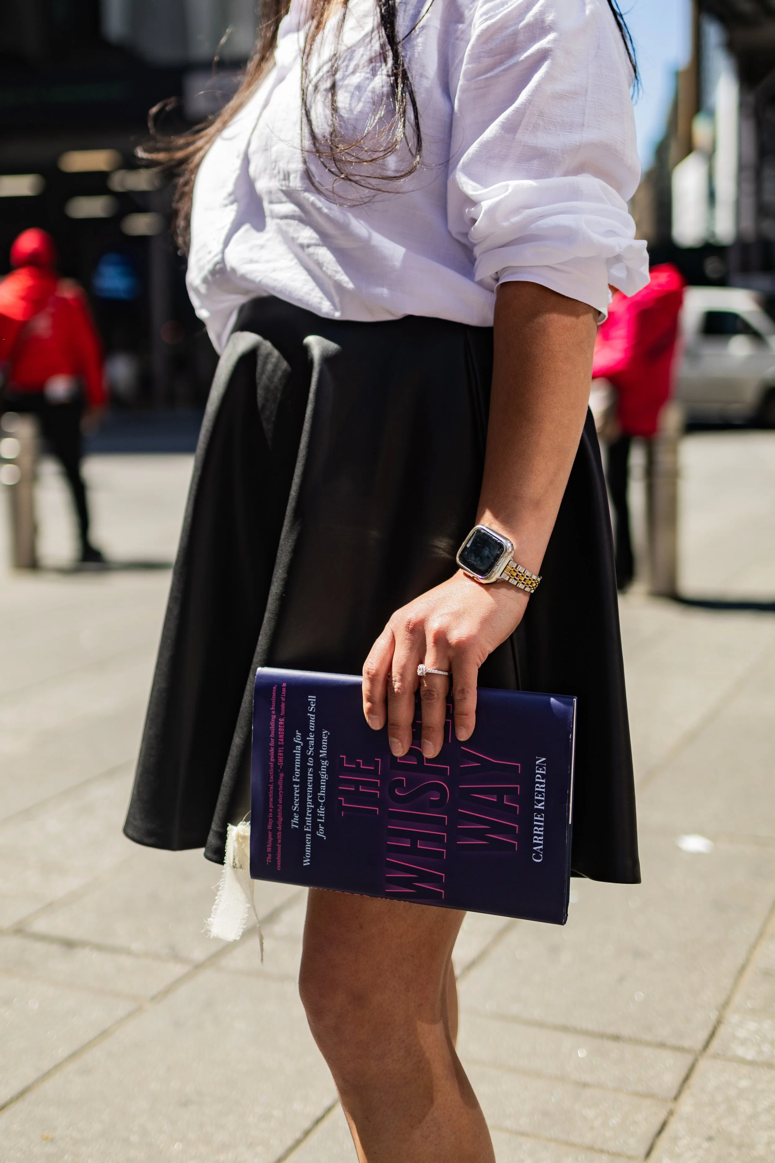 A woman wearing a white shirt with rolled-up sleeves, a black skirt, and a silver smartwatch, holding a book titled 'WOMEN WHO HUSTLE' on a city street.