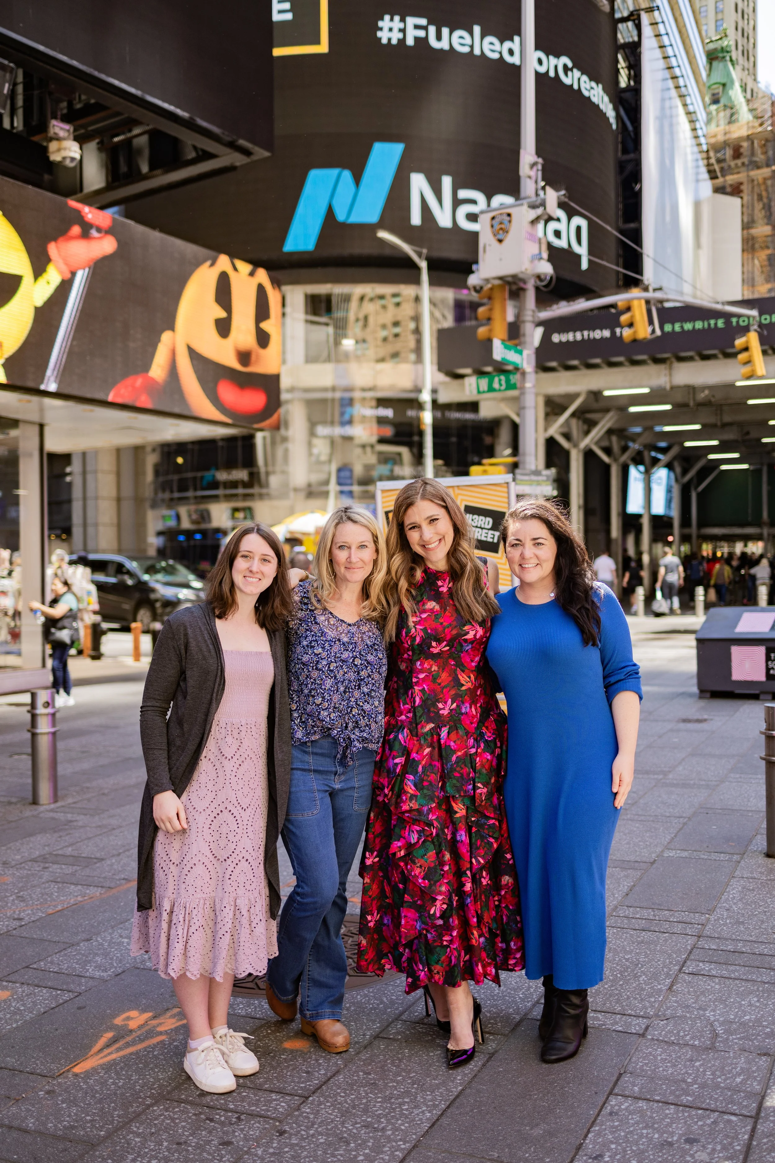 Four women standing on a city sidewalk in Times Square, New York City, smiling at the camera with digital billboards in the background.