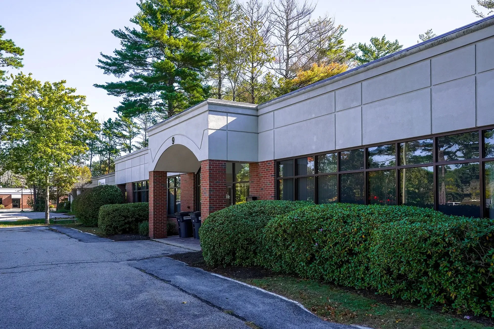 A commercial building with large windows and red brick columns, surrounded by green bushes and trees, with a parking lot in front.