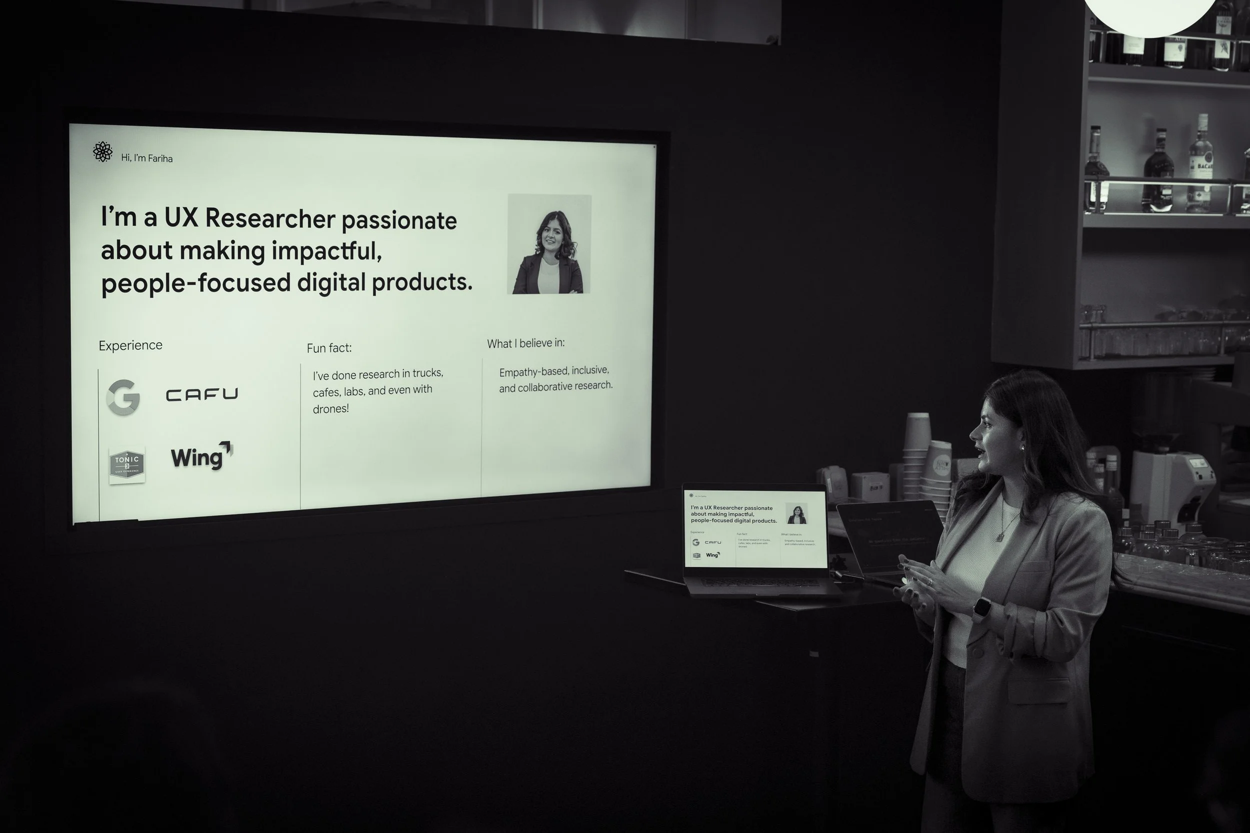 A woman standing at a counter with laptops and a large screen behind her, displaying a presentation about a UX researcher named Fariha.
