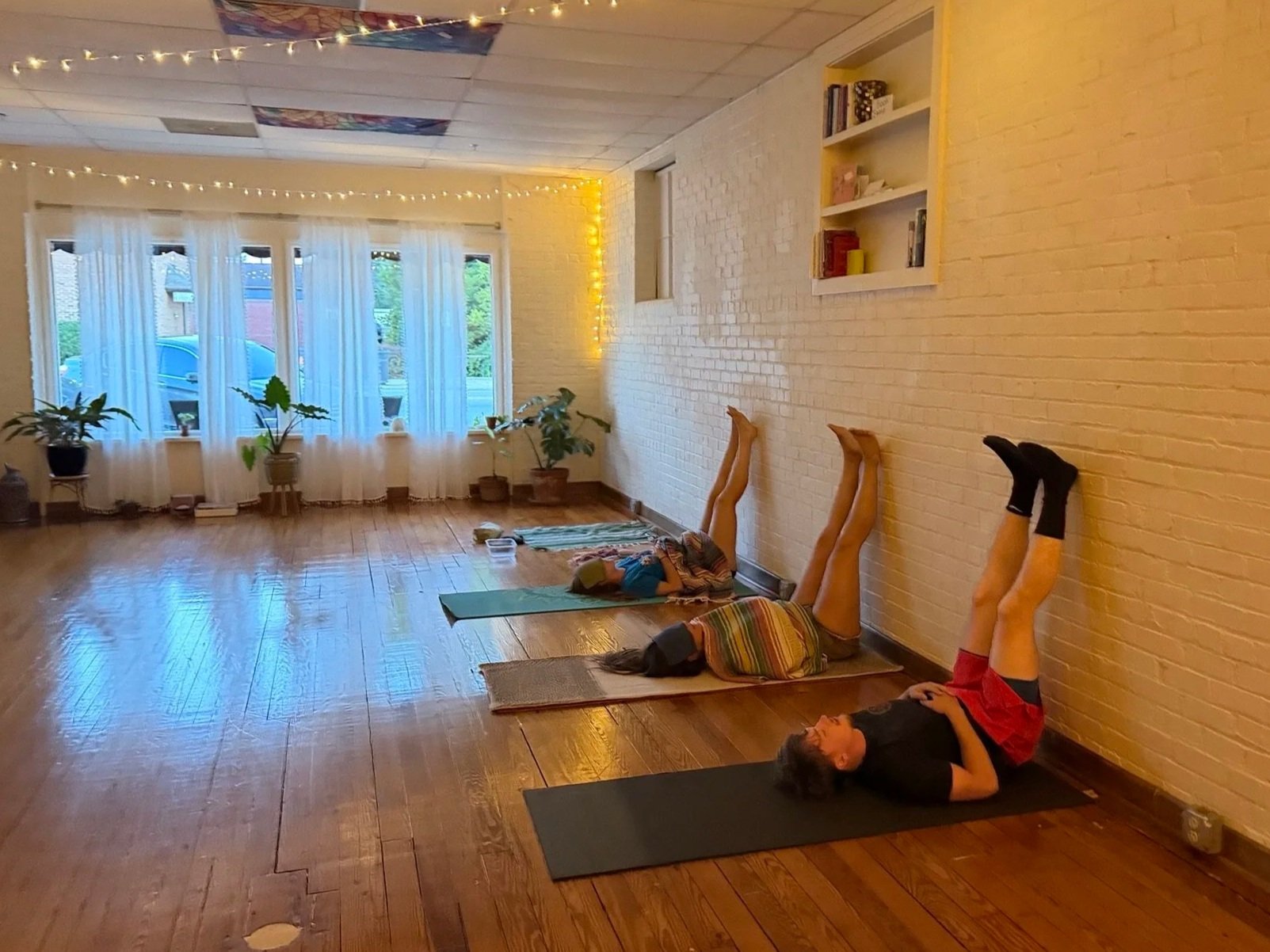 People lying on yoga mats with their legs up against a yellow brick wall during a yoga or relaxation class in a sunny room with white curtains and potted plants.