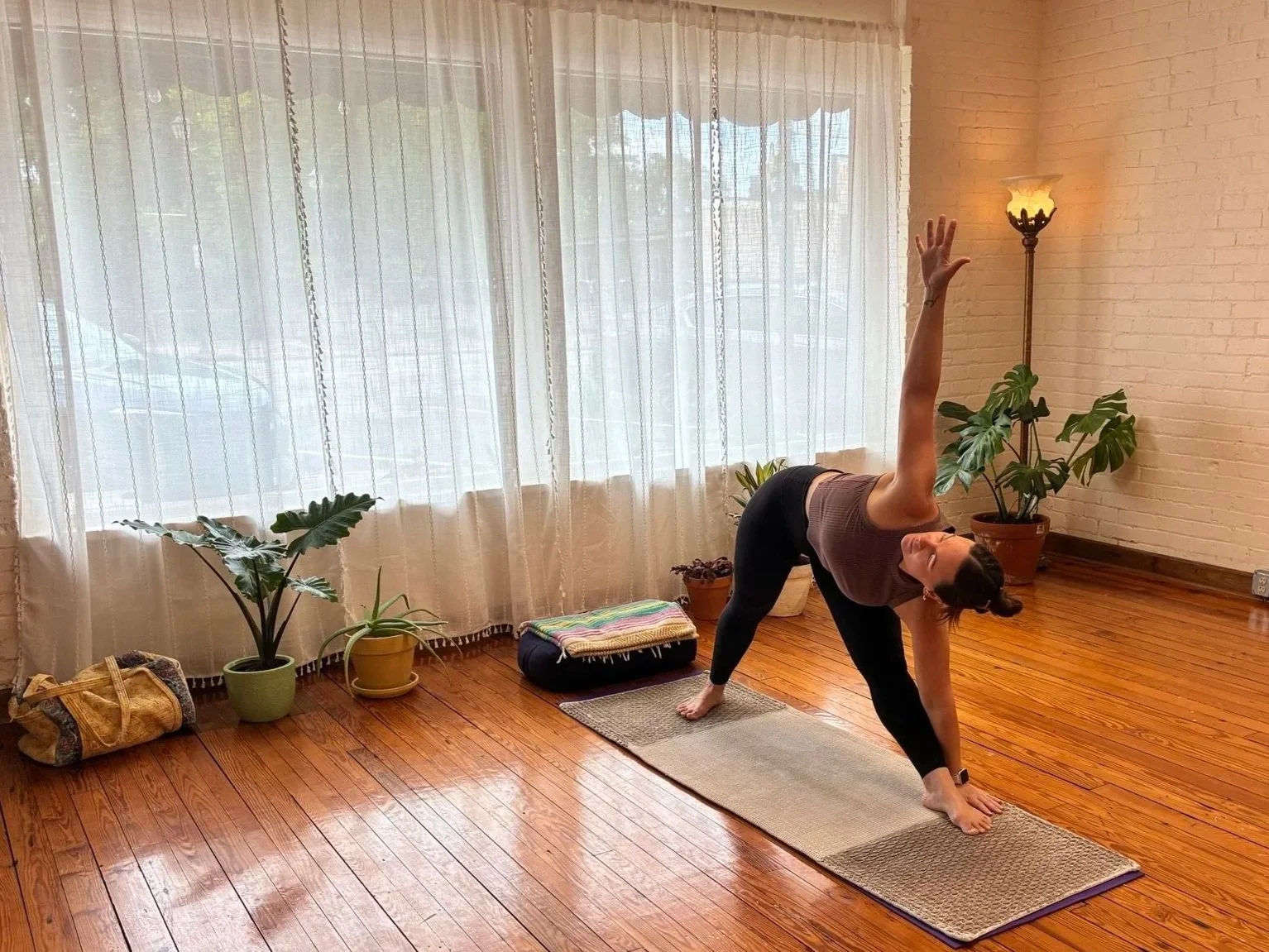 Founder, Melissa, practicing yoga in a room with wooden flooring, several potted plants, a window with sheer curtains, a standing lamp, and a small folded blanket on the floor.
