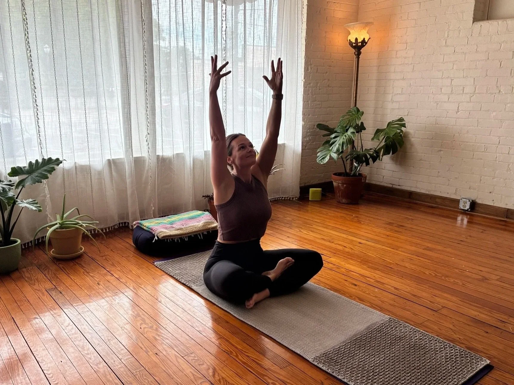 A woman practicing yoga in a well-lit room with hardwood floors, sitting cross-legged on a yoga mat, reaching upward with both arms extended, surrounded by potted plants and a floor lamp.