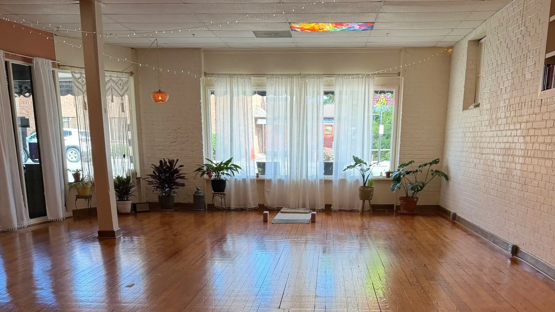Bright room with hardwood floor, white brick walls, large windows with sheer white curtains, potted plants, and string lights