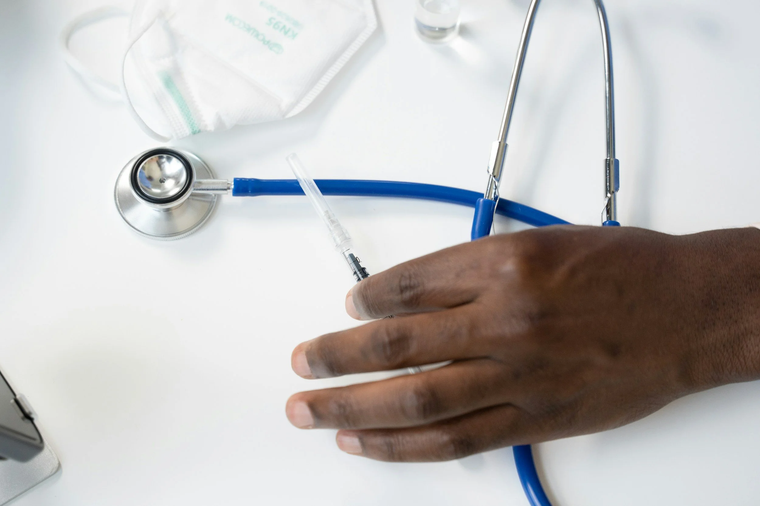 A healthcare professional's hand holding a syringe with a needle, surrounded by medical equipment including a stethoscope, white face mask, and medical tools on a white surface.