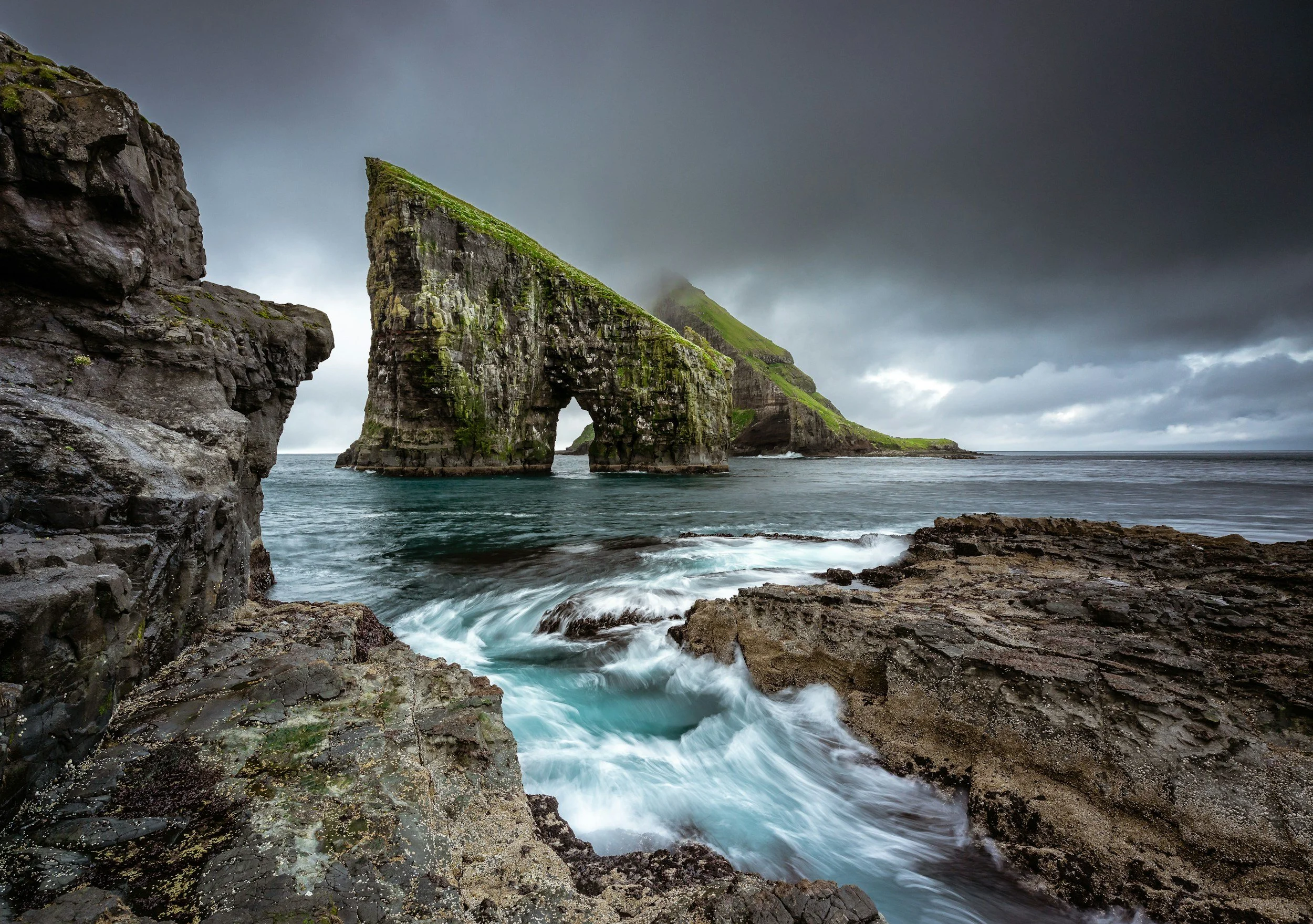 Squarespace design for entrepreneurs. A coastal landscape with large moss-covered rock formations, including a prominent arch-shaped sea stack, surrounded by water and dark, cloudy skies.