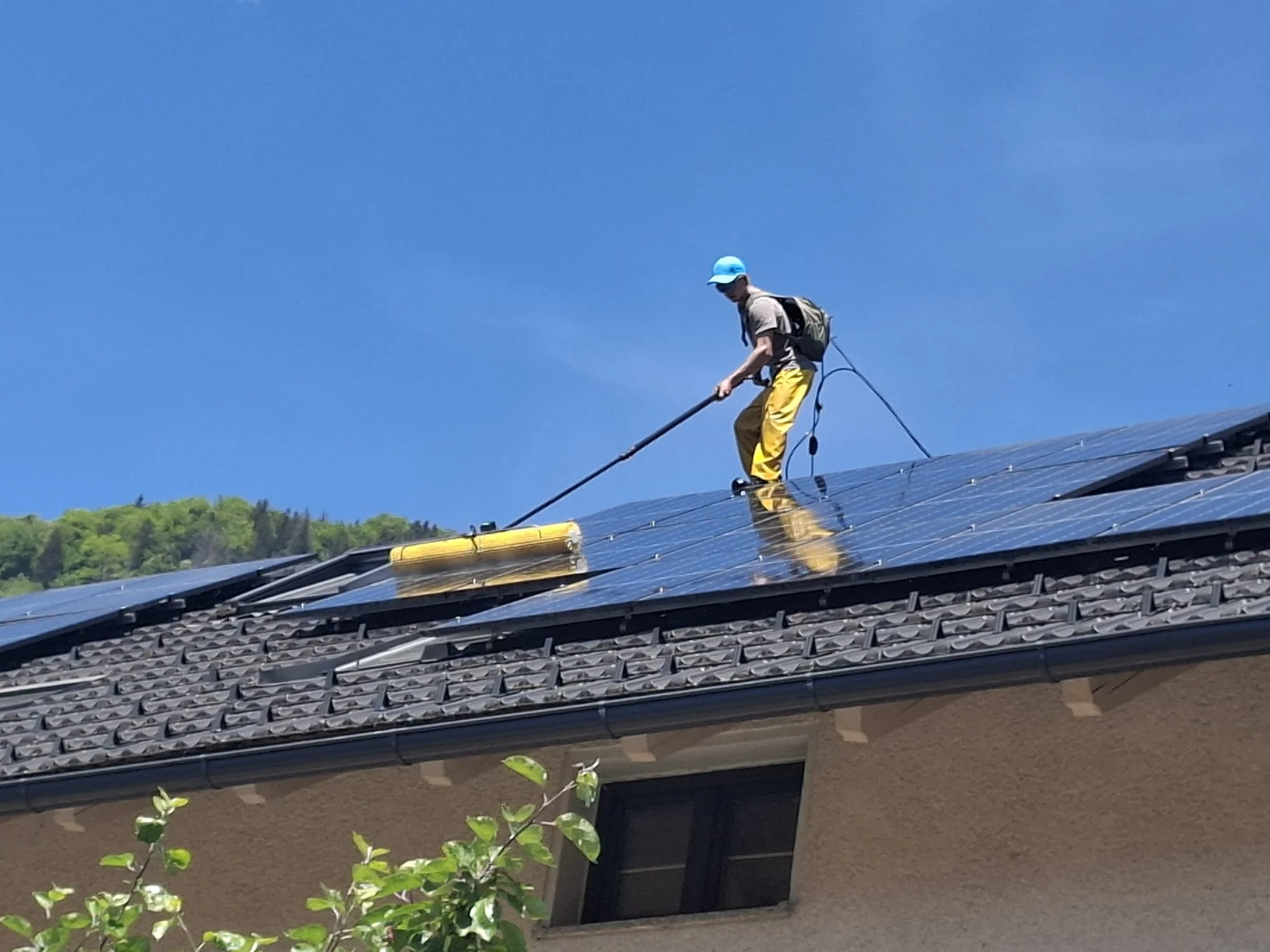 A person is on a rooftop installing solar panels, wearing a white cap, beige shirt, yellow pants, and a backpack, against a clear blue sky and green hills in the background.