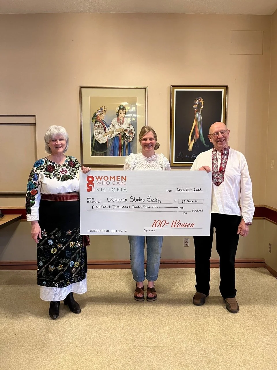 Three people are holding a large check in a room with beige walls and artwork. All three are smiling. The person on the left is a woman wearing a black skirt with colorful embroidery and a white blouse with floral patterns. The person in the middle is a woman with blonde hair wearing jeans and a white lacy top. The person on the right is a man with glasses, wearing a white shirt with red embroidery. The check is addressed to Ukrainian Studies Society for $18,300, dated April 20th, 2022.