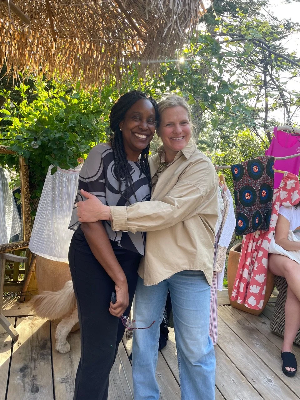 Two women hugging and smiling on a wooden deck outdoors. One woman has curly hair and is wearing a gray and black patterned top, while the other has blonde hair and is wearing a beige shirt and jeans. There are plants, trees, and colorful fabrics in the background.