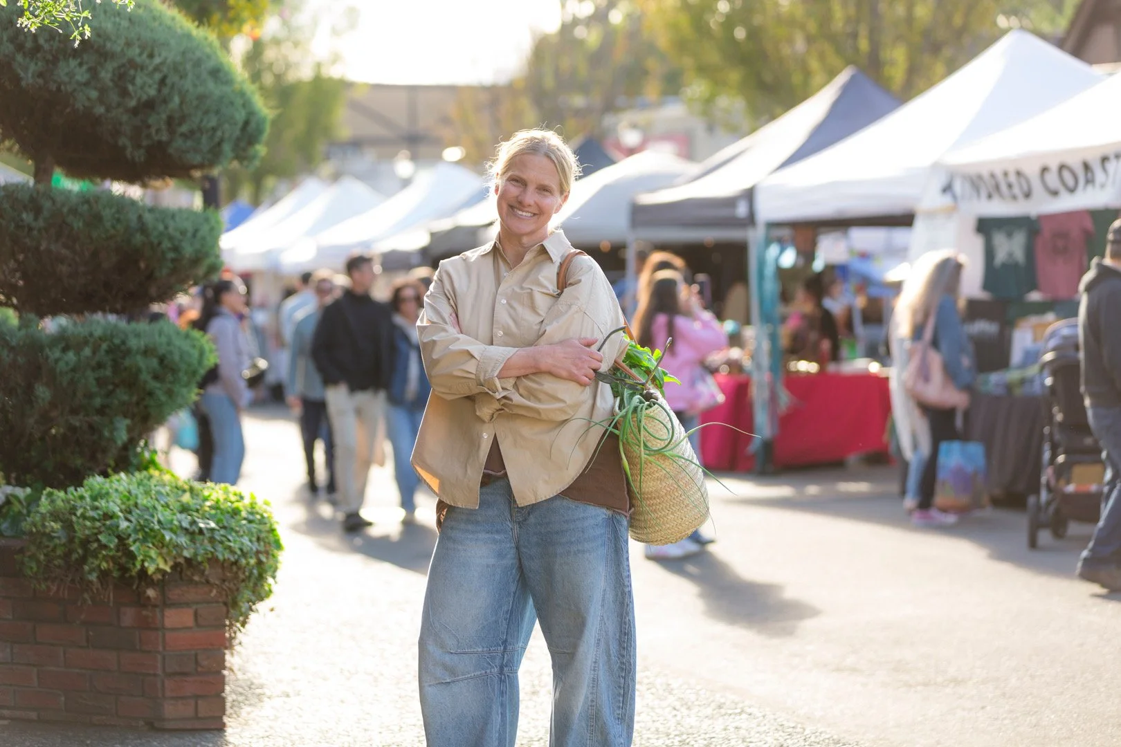 A woman standing at an outdoor market, smiling, holding a bag of green vegetables, with vendor tents and shoppers in the background.
