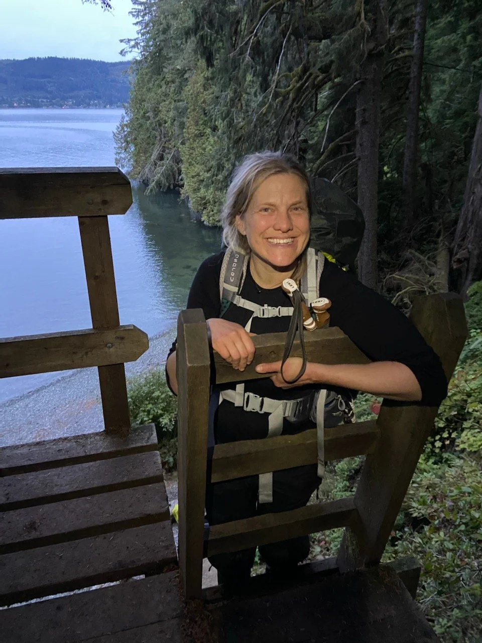A smiling woman with a backpack leaning on a wooden railing on a forest trail near a lake, with trees and mountains in the background.