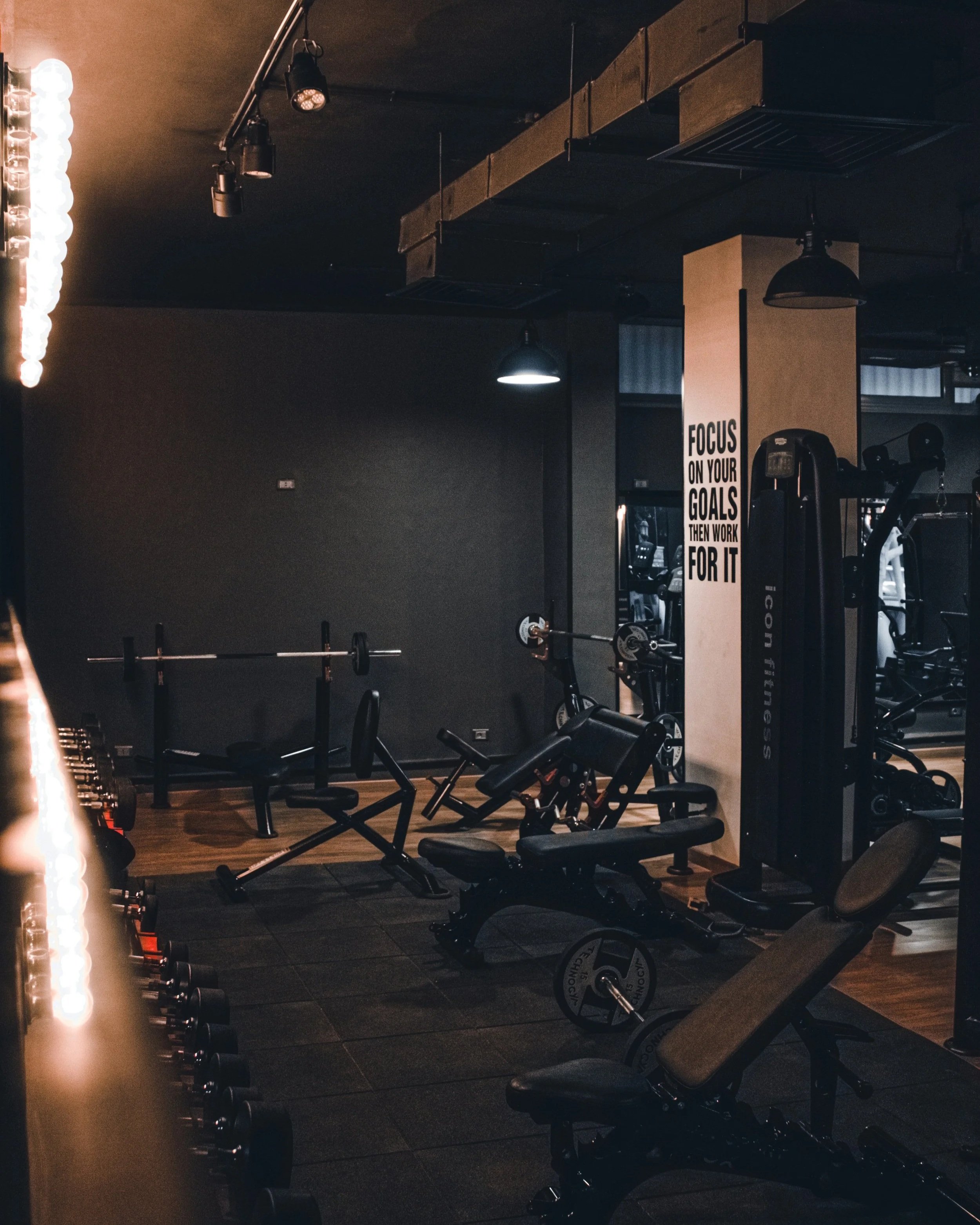 A dimly lit gym with workout equipment including a barbell on a rack, exercise benches, weights, and cardio equipment. A motivational poster on the wall reads, "Focus on your goals then work for it."