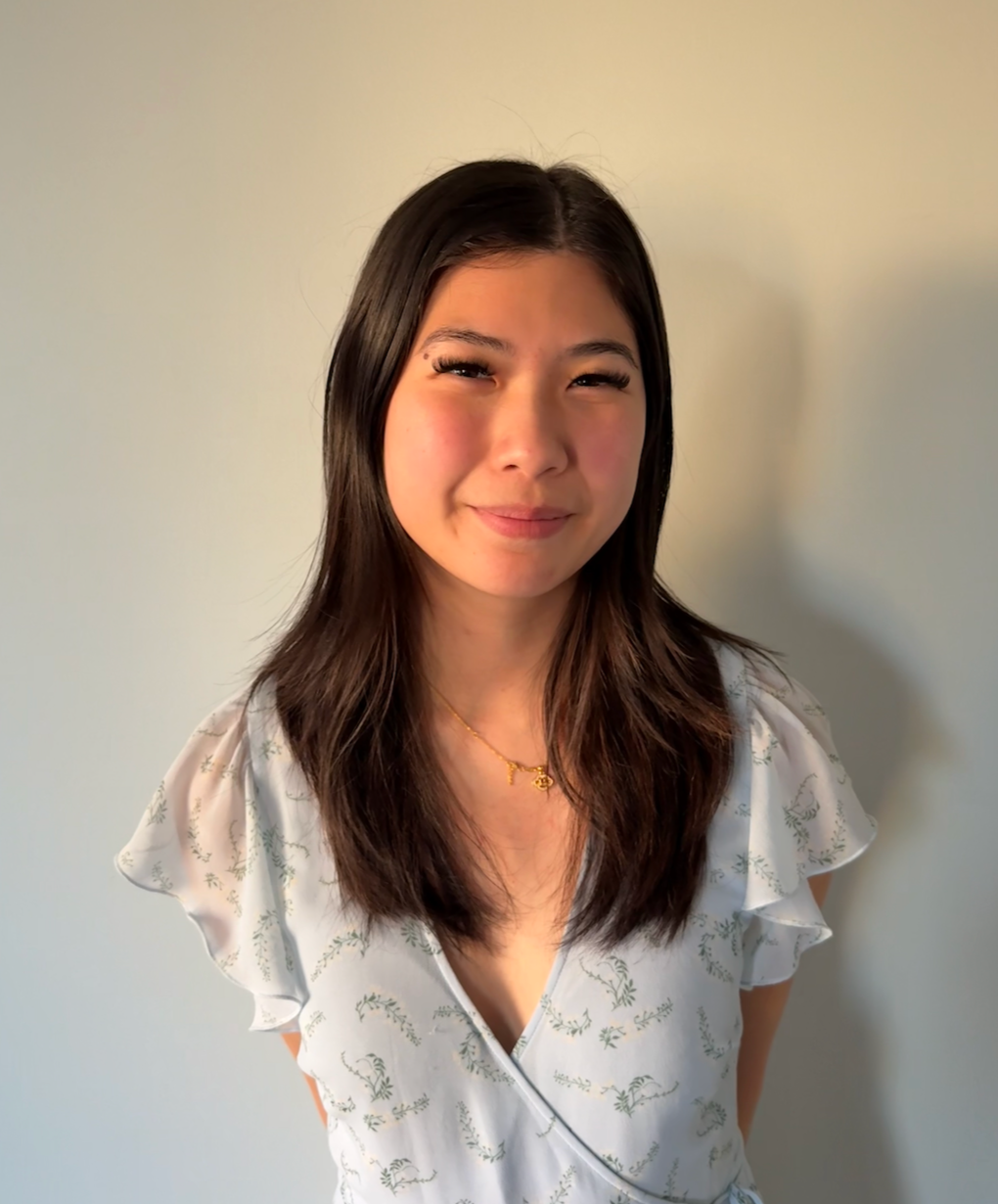 A young woman with long dark hair smiling, wearing a white top with a nature-inspired pattern and a delicate gold necklace, standing against a plain light-colored wall. A kinesiologist graduating from University of British Columbia, founder