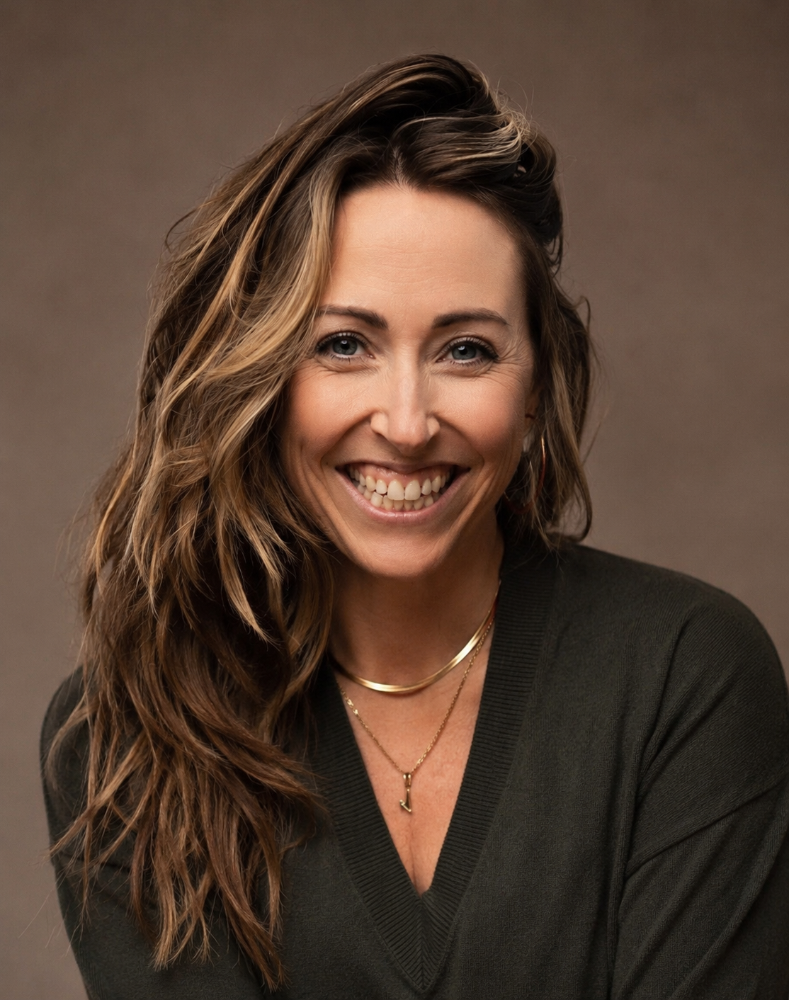 A woman with wavy brown hair, smiling, wearing a black top and layered gold necklaces, posing against a neutral background.