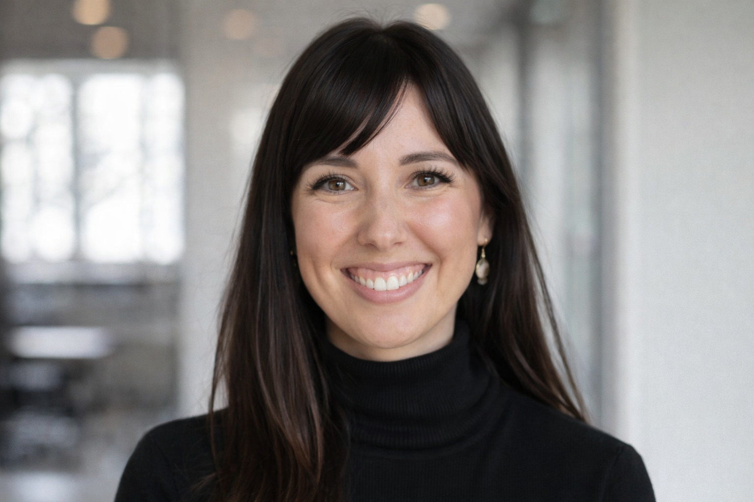 A woman with long dark hair and light brown eyes smiling, wearing a black turtleneck and pearl earrings, in a modern indoor setting with blurred windows in the background.