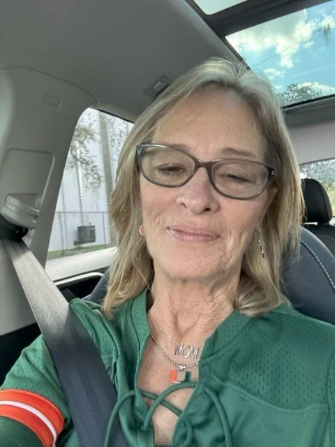 A woman with blonde hair, glasses, and a green sports jersey, sitting in a car and smiling with one eye closed.