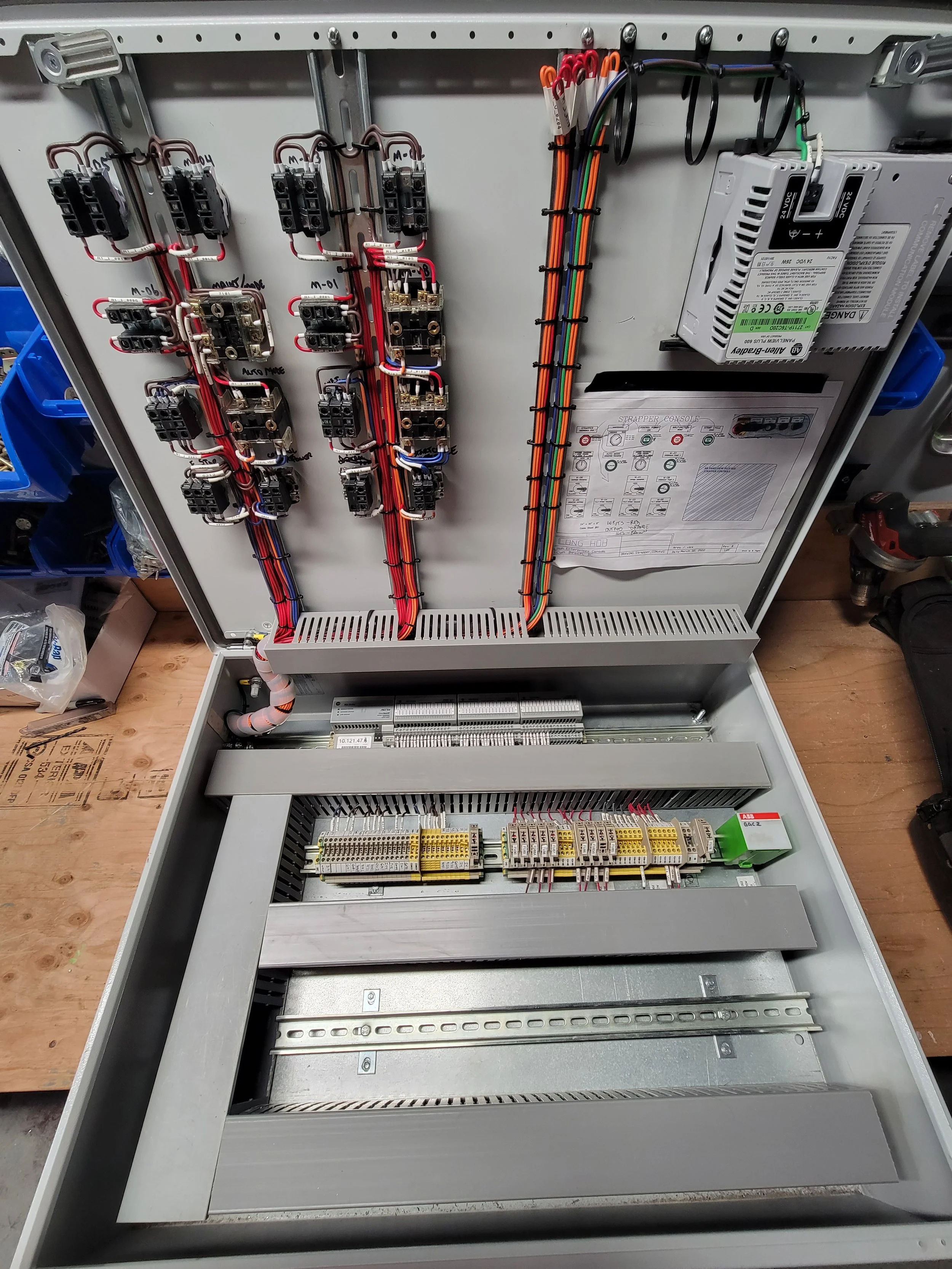 Open electrical control panel with various wiring, relays, power supply, and terminal blocks, with detailed wiring diagrams attached inside the panel door.