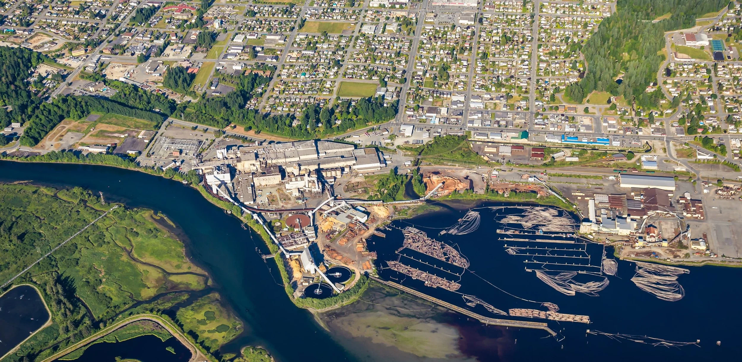 Aerial view of a city with a river running through it, showing a mix of residential neighborhoods, industrial areas, and green spaces, with boats docked along the waterfront.