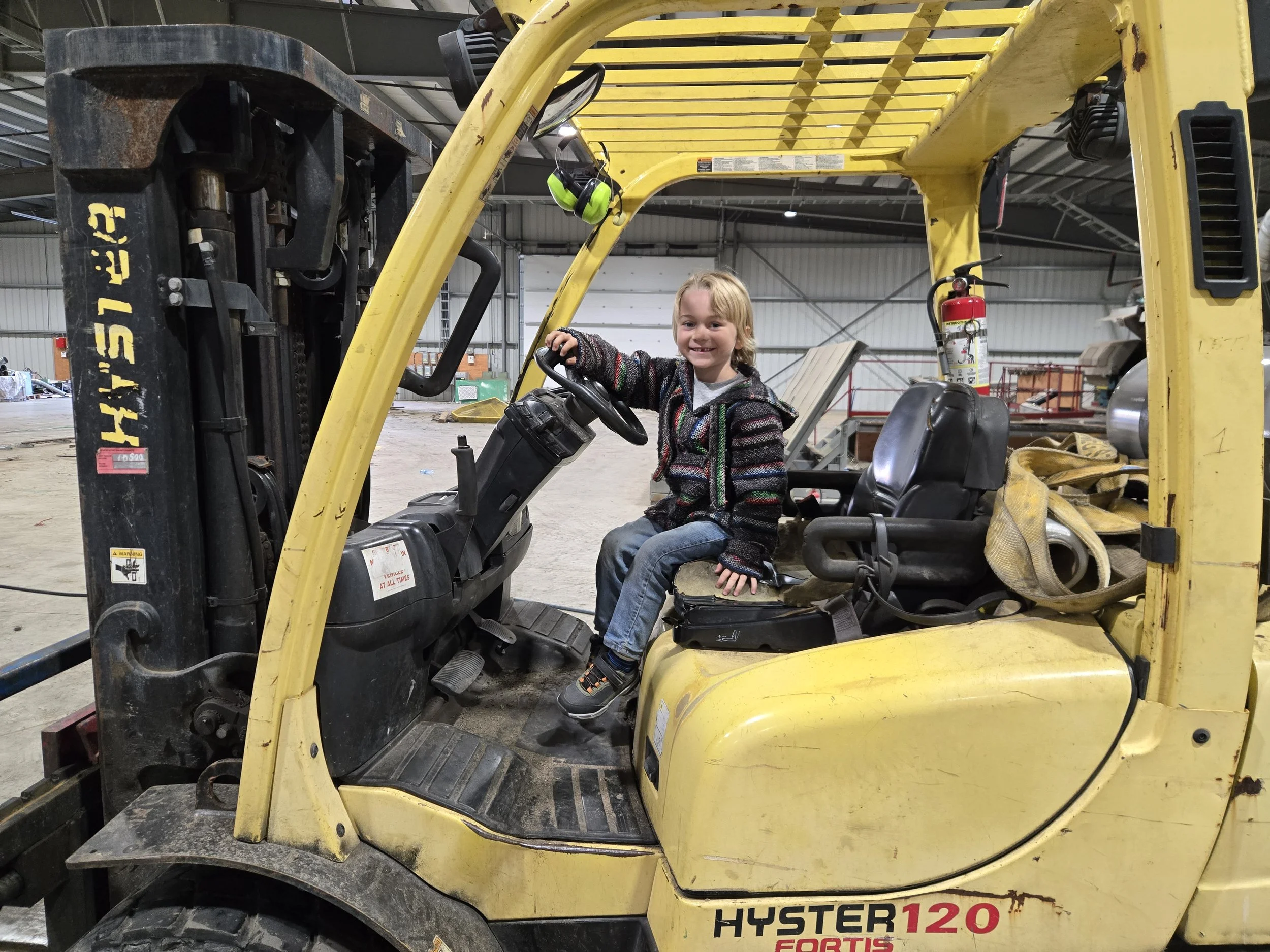 A young girl with blonde hair, wearing a colorful sweater and jeans, sitting in the driver's seat of a yellow forklift inside a large industrial warehouse.