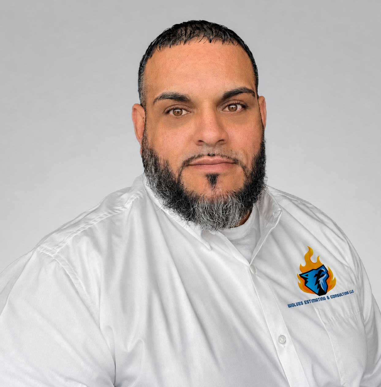 Portrait of a man with short dark hair, beard, and mustache, wearing a white shirt with a logo on the chest that reads 'Wolves Estimating & Consulting LLC' with a wolf head and flames. The background is plain light gray.