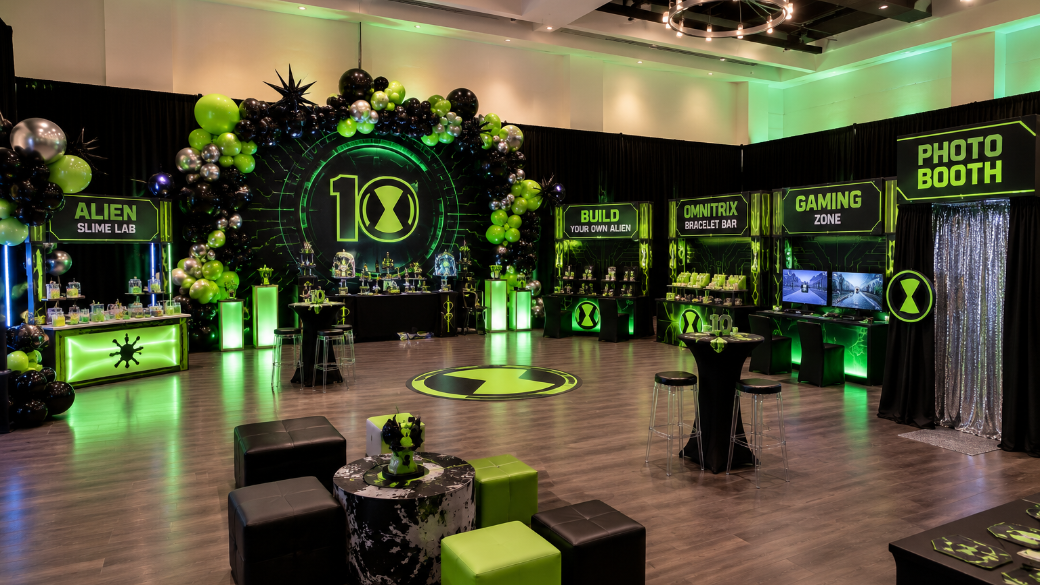 A decorated event space with a gaming theme, featuring green and black balloons, neon green lighting, and various activity stations including an alien slime lab, build your own alien, Omnitrix bracelet bar, gaming zone, and photo booth.