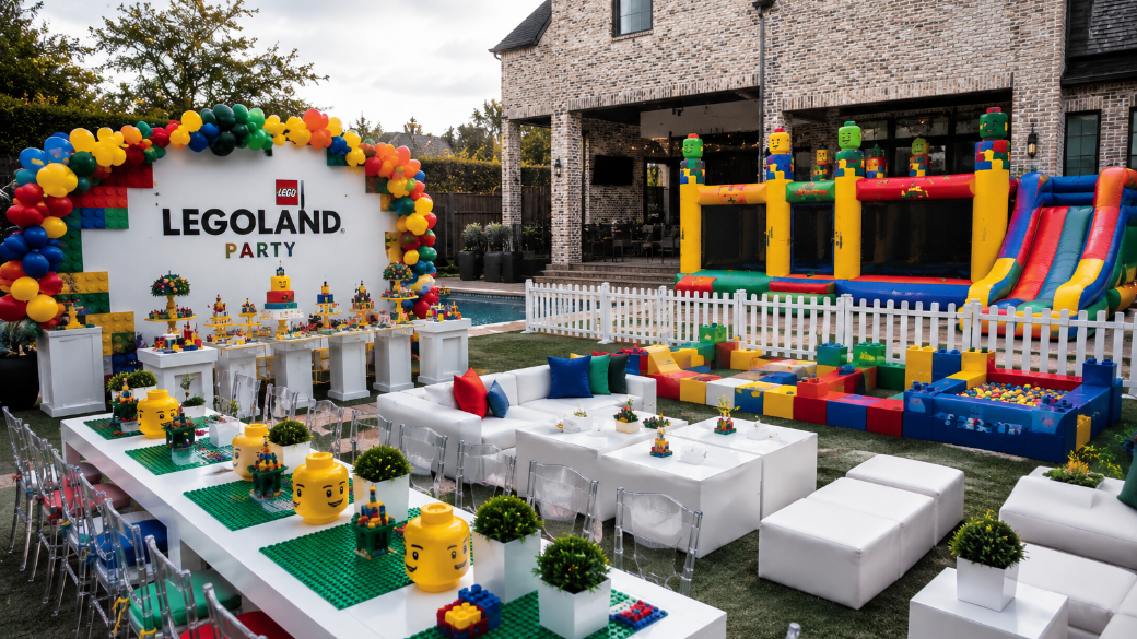 Backyard LEGO-themed party setup with a colorful balloon arch, LEGO decorations, a LEGO brick wall, LEGO-inspired seating, and an inflatable LEGO castle and slide.