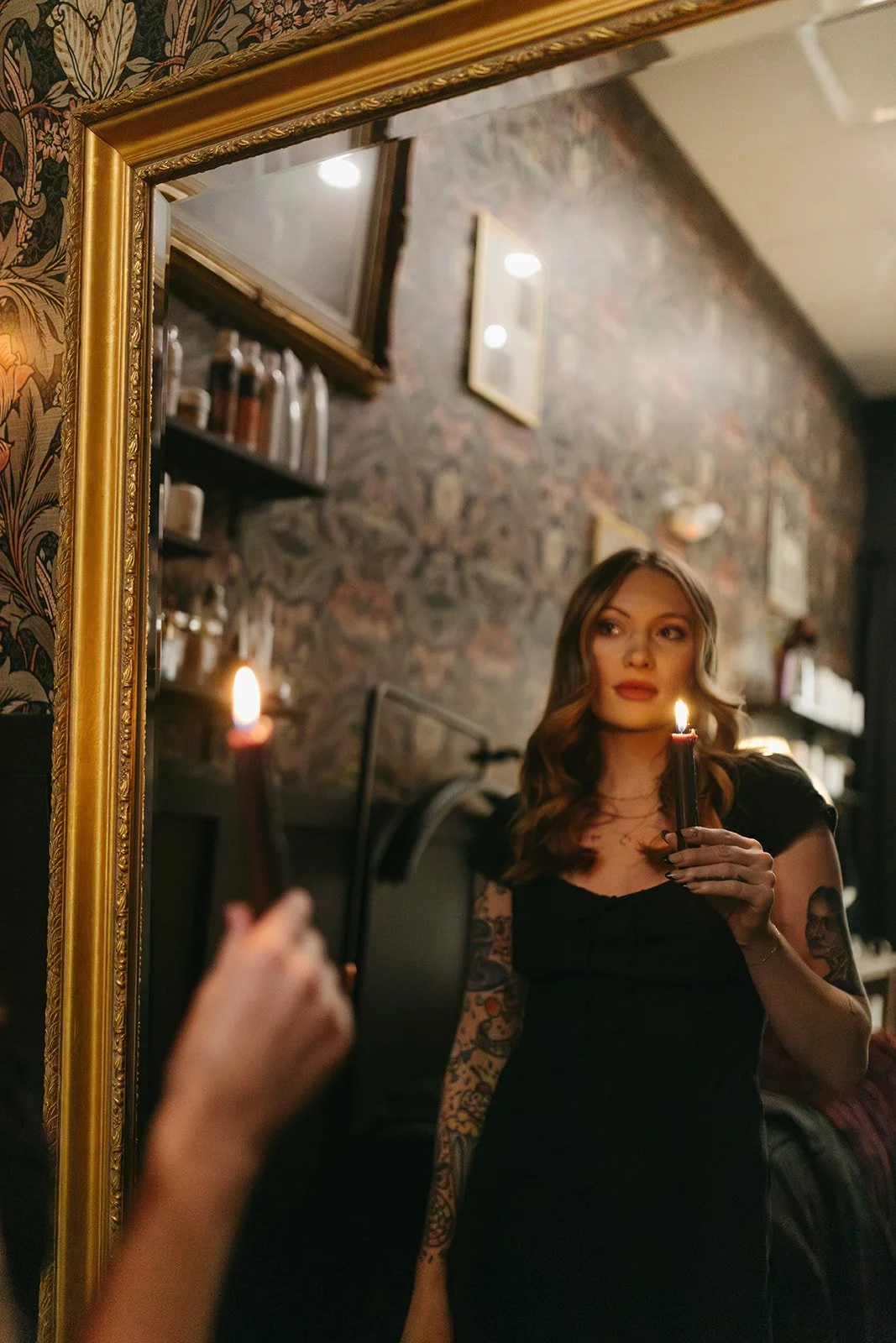 A woman with wavy hair and tattoos on her arms is holding a lit candle in front of a mirror, creating a reflection of herself. The room has a rustic brick wall with shelves and bottles, and the woman is wearing a black top.