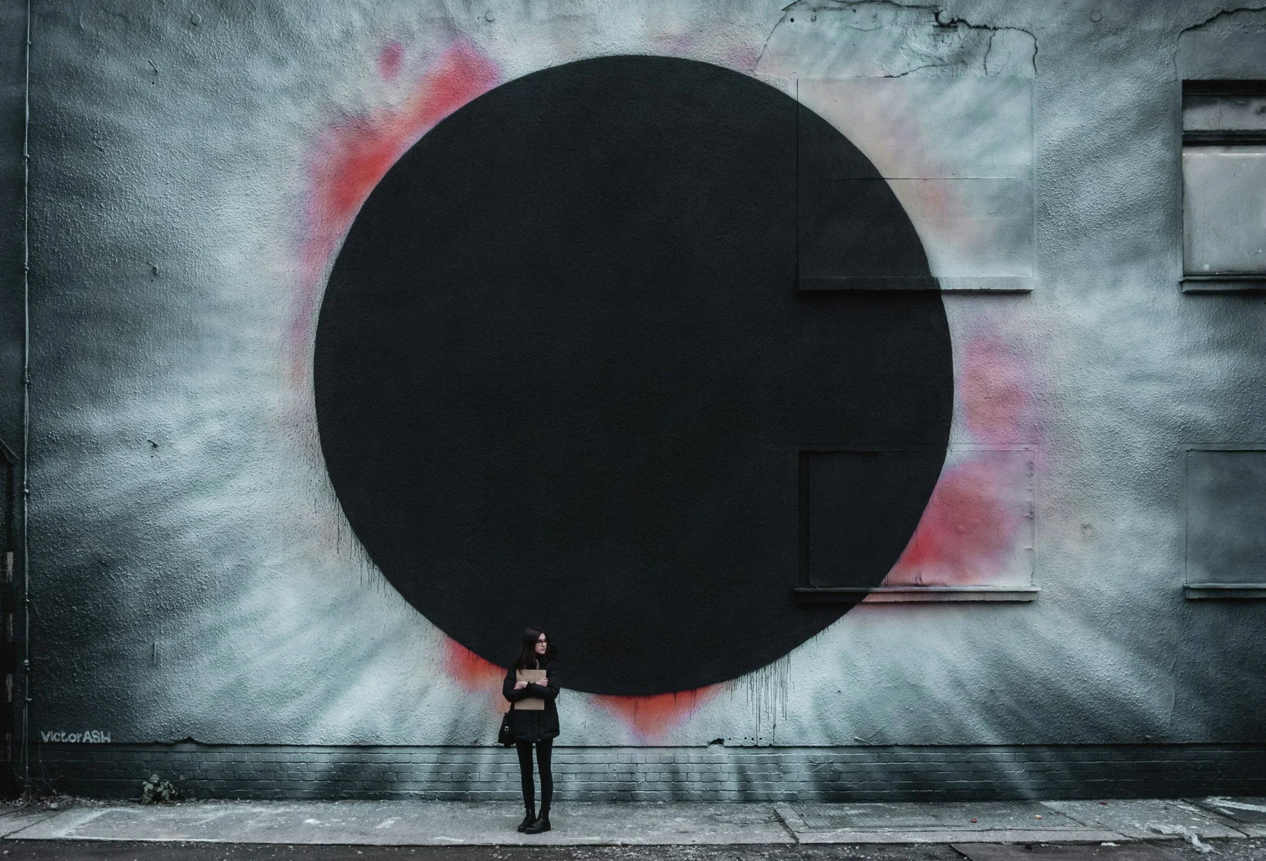 A woman standing against a graffiti mural of a large black circle on a concrete wall, with pink and gray spray paint accents. She is holding a folder and looking to her left.