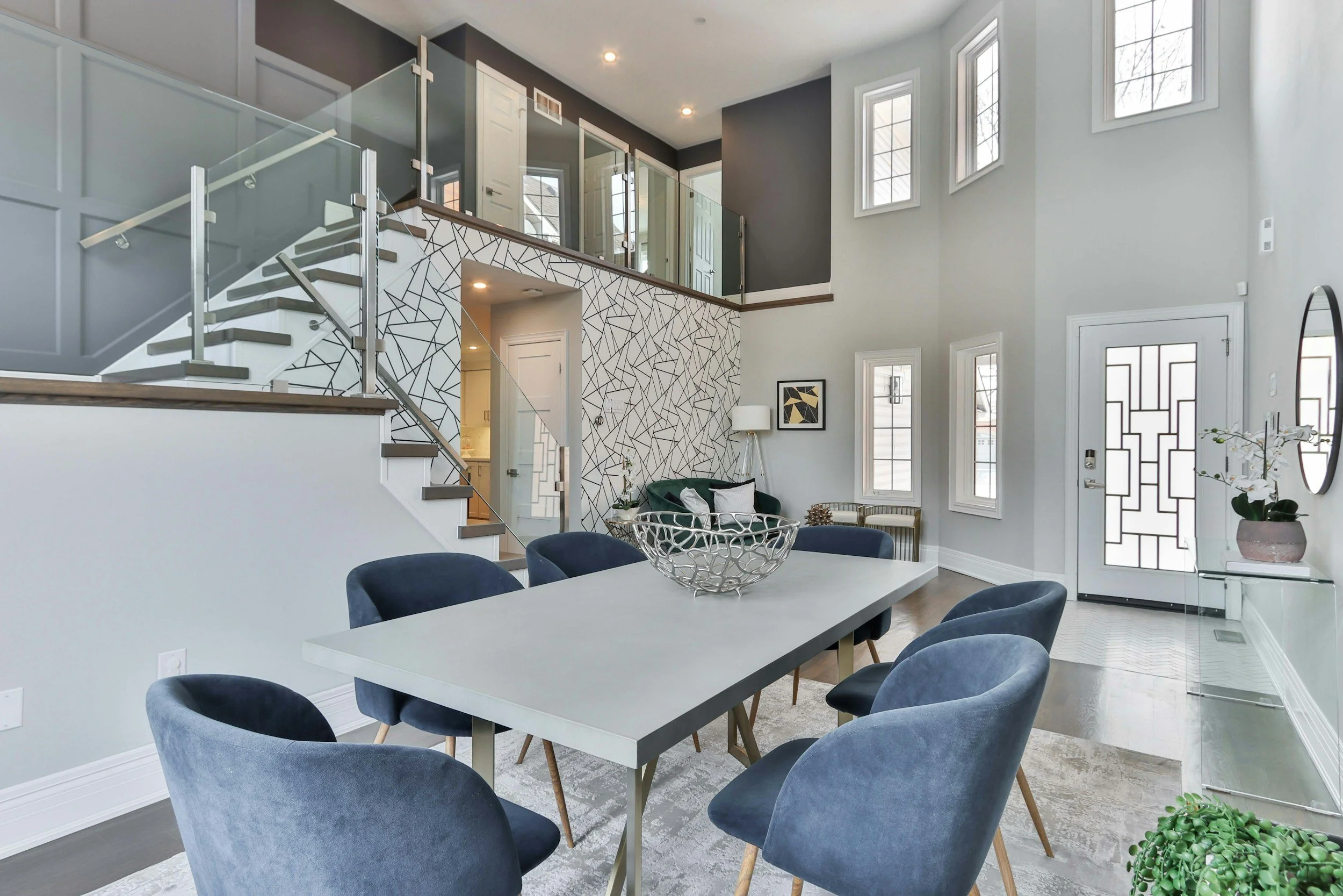 Modern living room with high ceilings, large windows, gray walls, a geometric accent wall, and a dining area with a white table and blue chairs.