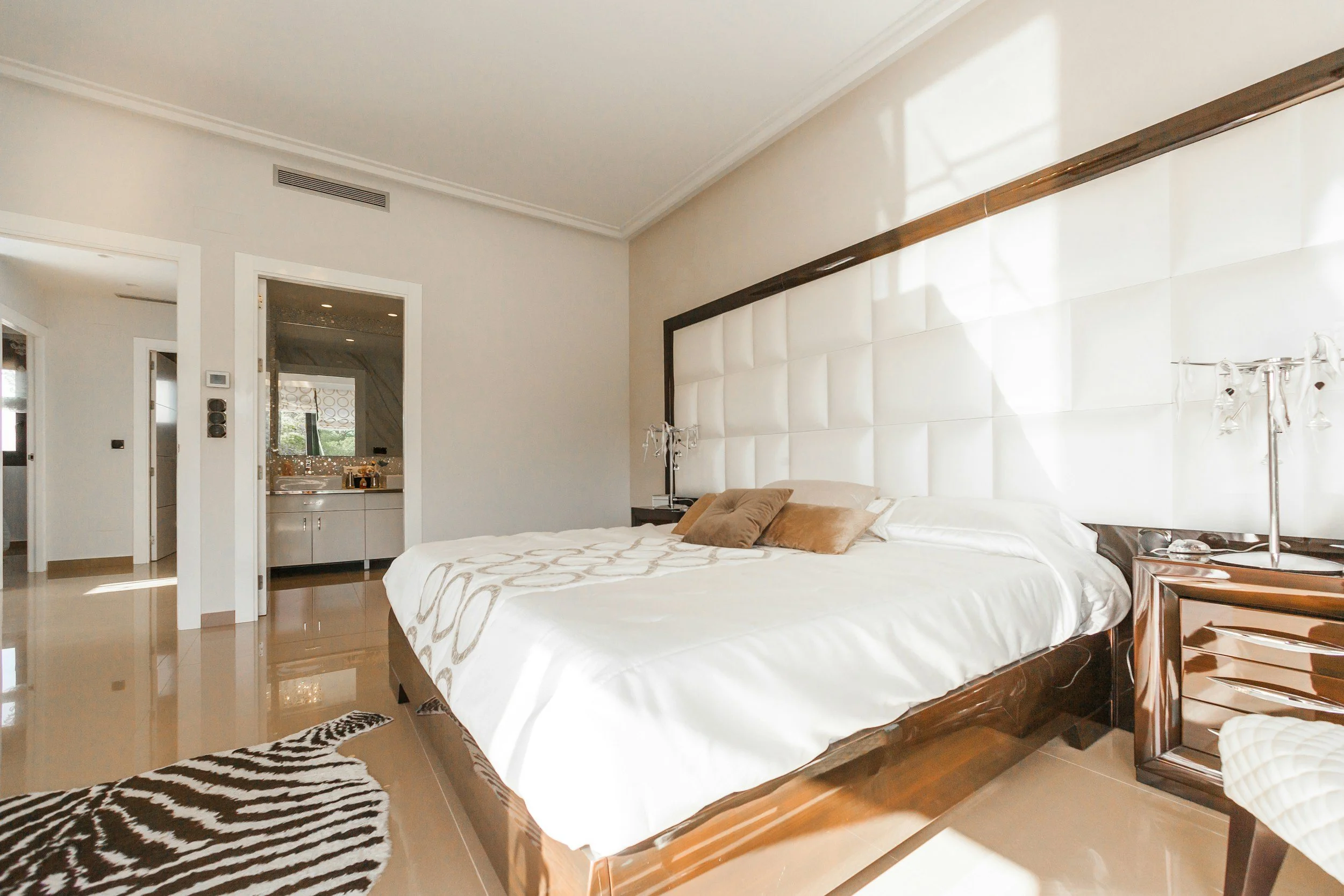 A bedroom with a large bed, decorated with beige pillows and a white and beige bedspread, next to a window with sunlight casting shadows on the wall. There is a zebra print rug on the wooden floor and a zebra print bench at the foot of the bed. A sid