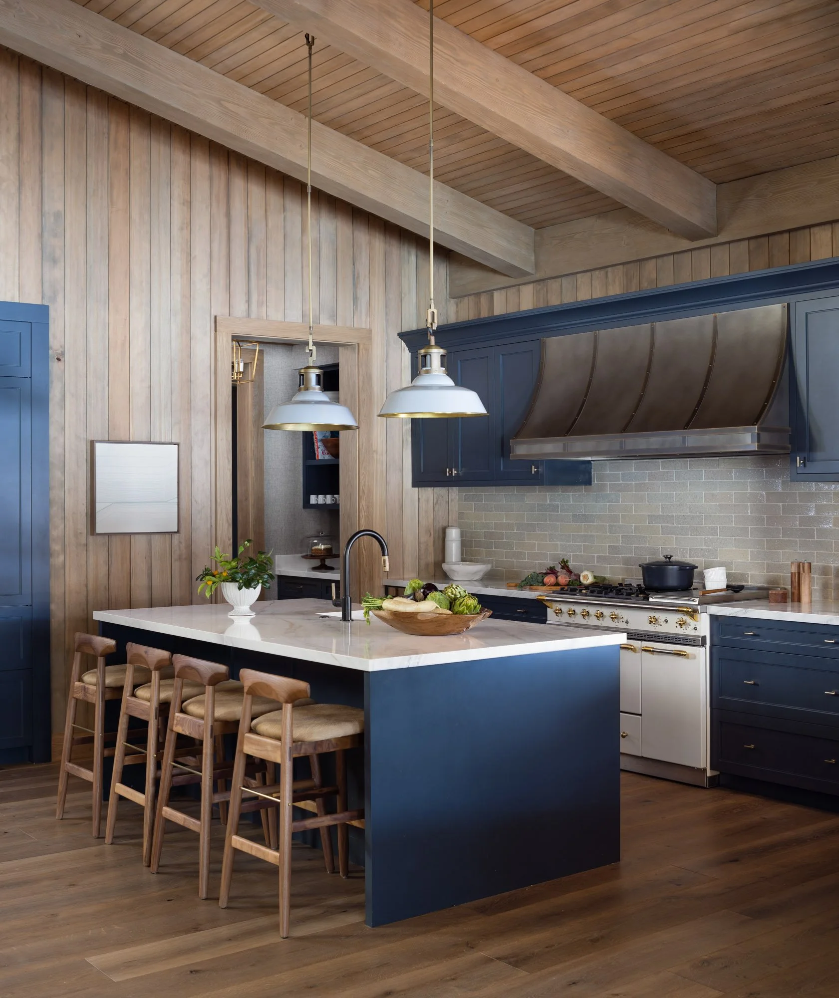 A modern kitchen with a white marble island counter and wooden bar stools. The kitchen has dark blue cabinets and a stainless steel range hood. The walls and ceiling are made of light-colored wooden panels, and there are two pendant lights hanging above the island.