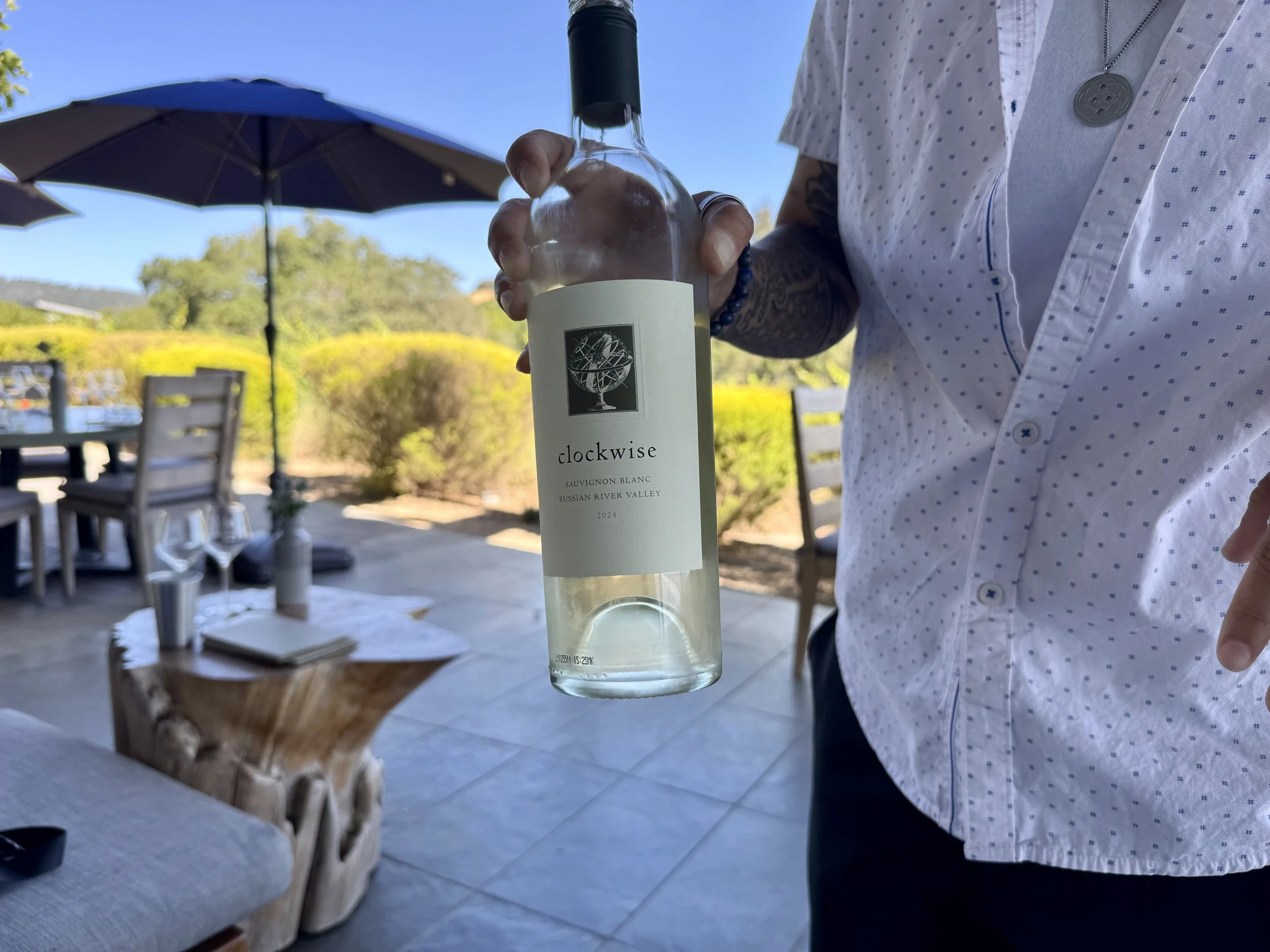 Person holding a bottle of white wine outdoors on a sunny day, with trees, from an angle focusing on the bottle and part of the person's torso.