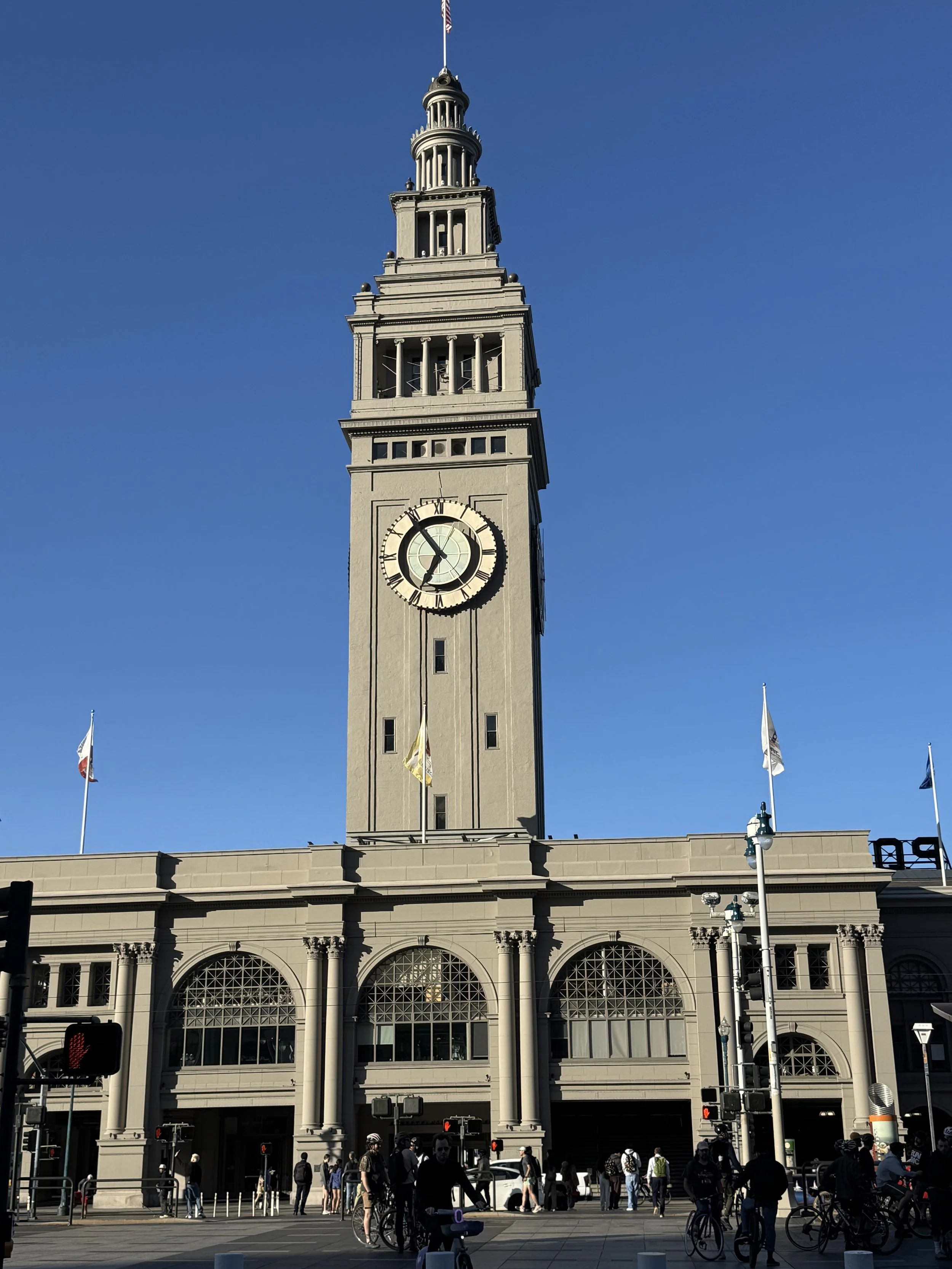 Ferry plaza building and food hall and historic Clock tower with a flag on top, part of a historic building with large arched windows, people walking and biking in front, American flags, and traffic lights.