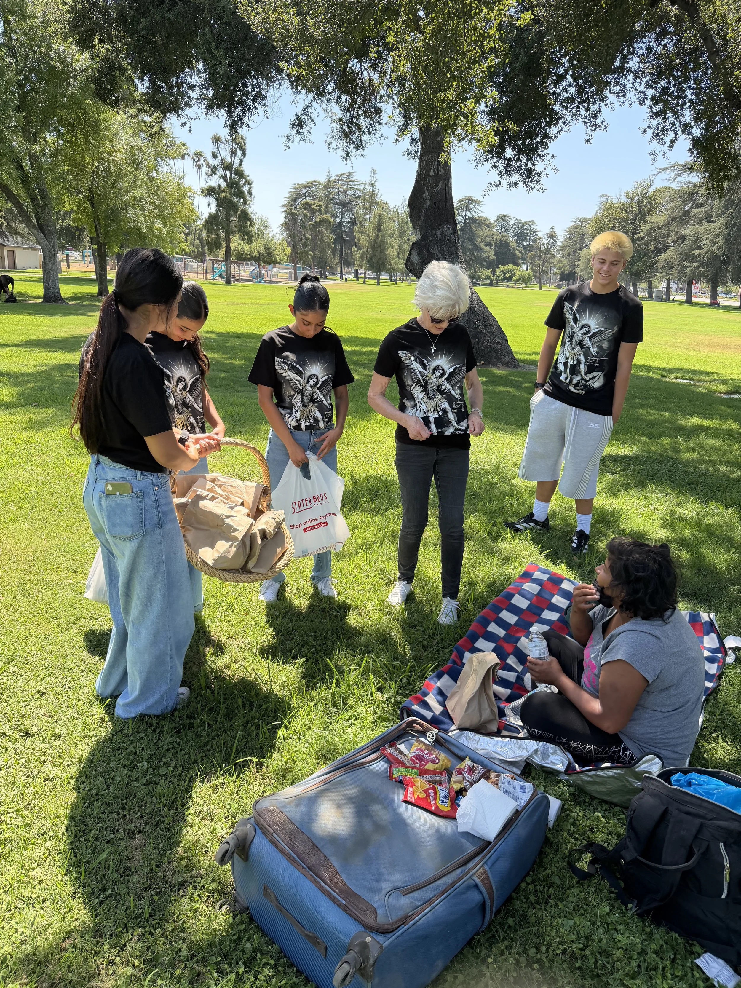 A group of five people, four women and one man, gathered outdoors under a large tree in a park, preparing for a picnic with bags and snacks on a blanket. One woman is sitting on the blanket holding a water bottle, while the others stand around looking at items and bags.