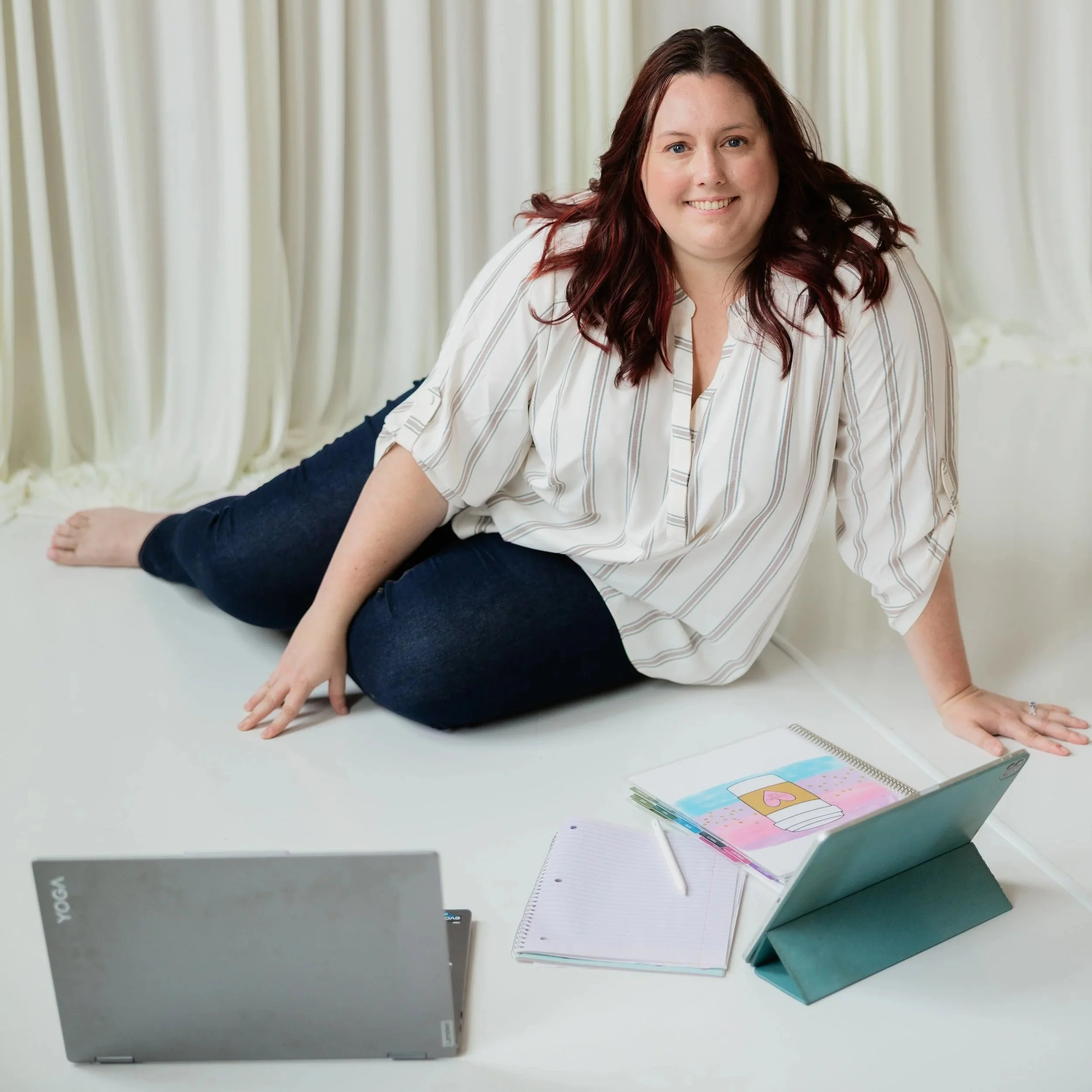 An open laptop with a gray cover and a white keyboard, a notebook with colorful pages and a spiral binding, a closed teal notebook, and a few loose papers on a white table, with a woman sitting behind them in front of cream-colored curtains.
