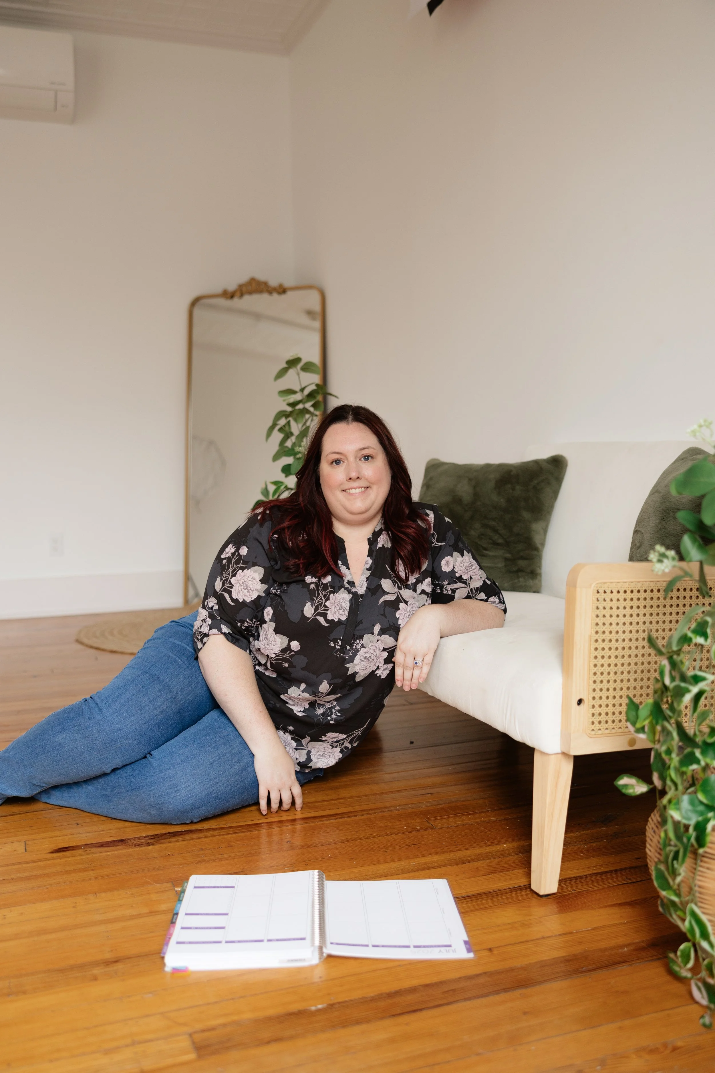 A woman with long reddish-brown hair and light skin, wearing a black floral blouse and blue jeans, reclining on the wooden floor of a room next to a white bed, with green cushions, a cactus, and a mirror in the background.