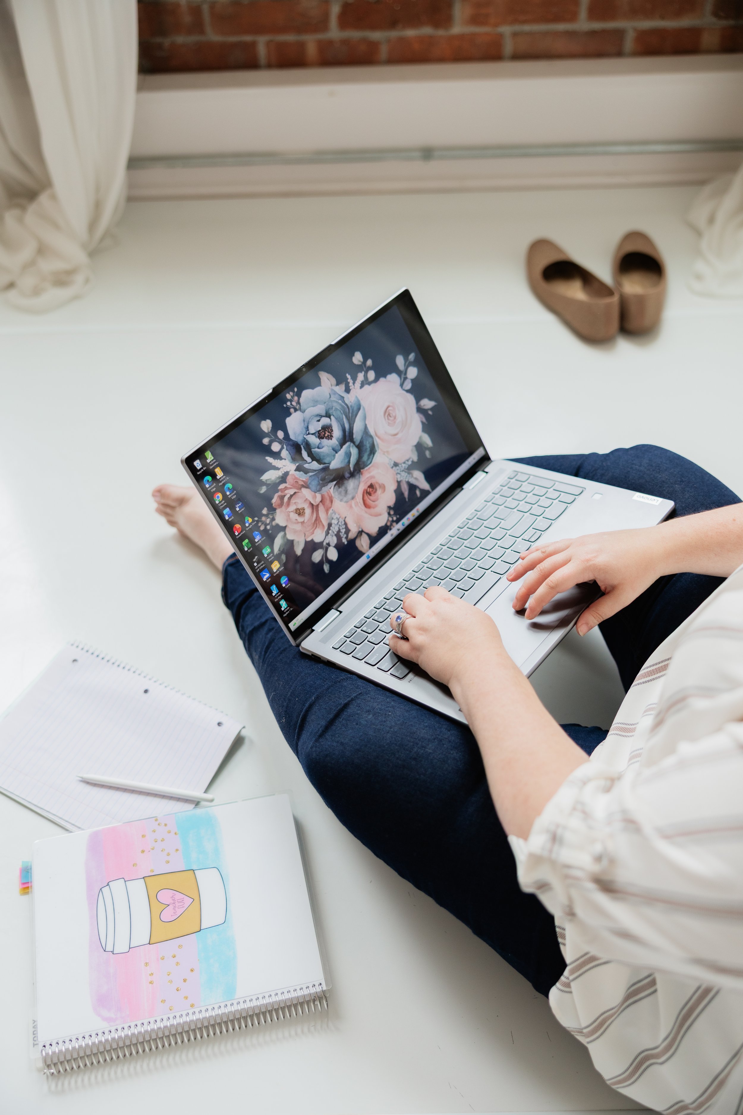 A person sitting on a white surface using a laptop with a floral wallpaper on the screen. There are notebooks and a colorful illustration of a coffee cup on the table. Shoes are placed on the floor in the background near a brick wall and window with curtains.
