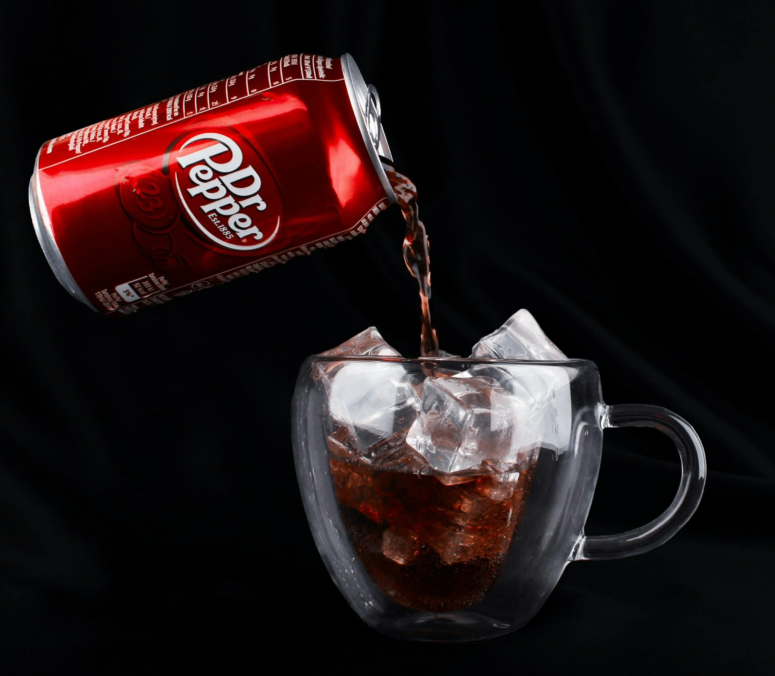 A can of Dr. Pepper soda tipped over pouring into a glass filled with ice, with a black background.