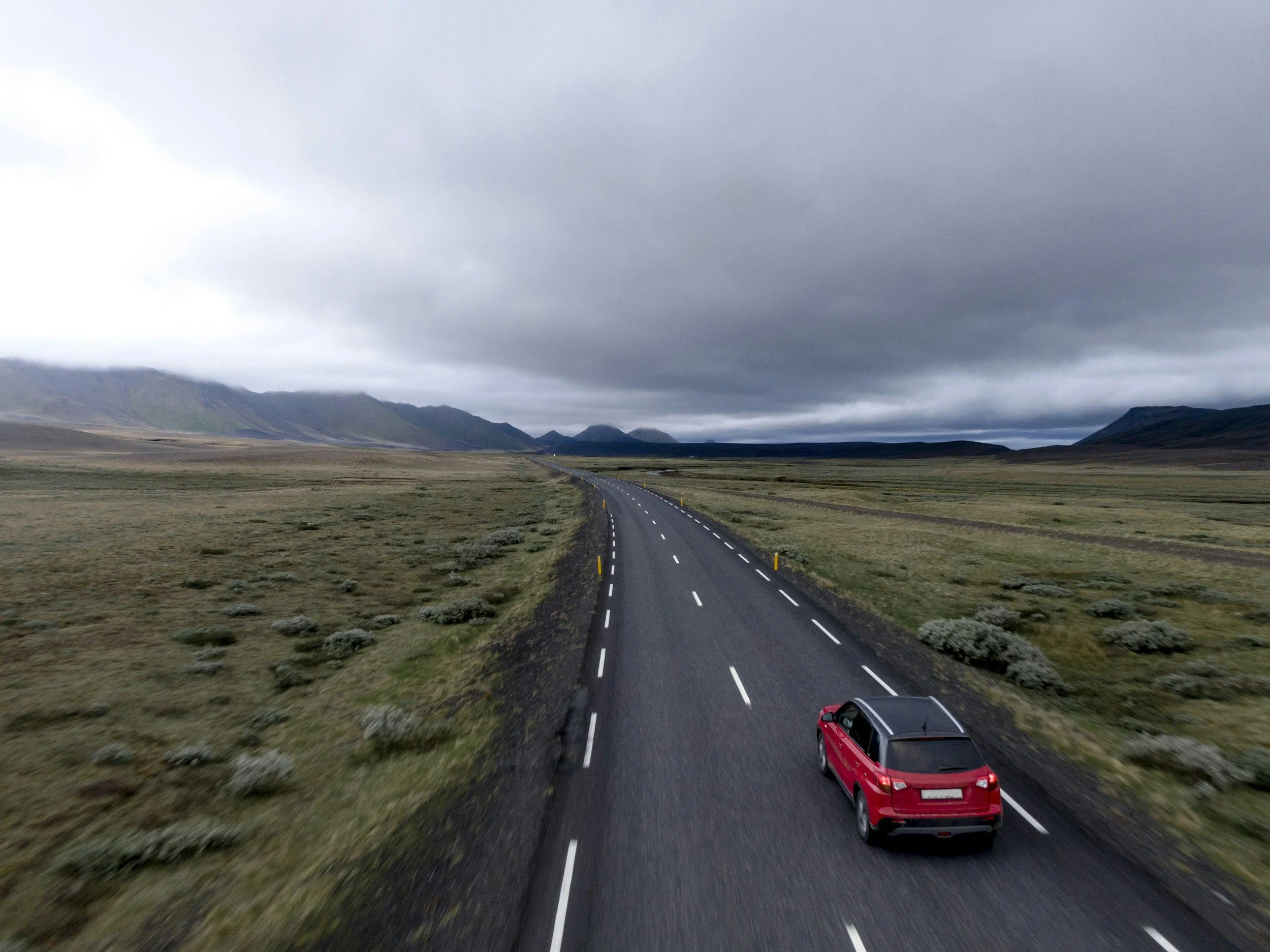 A red car driving on a long, straight road through a mountainous landscape under a cloudy sky.