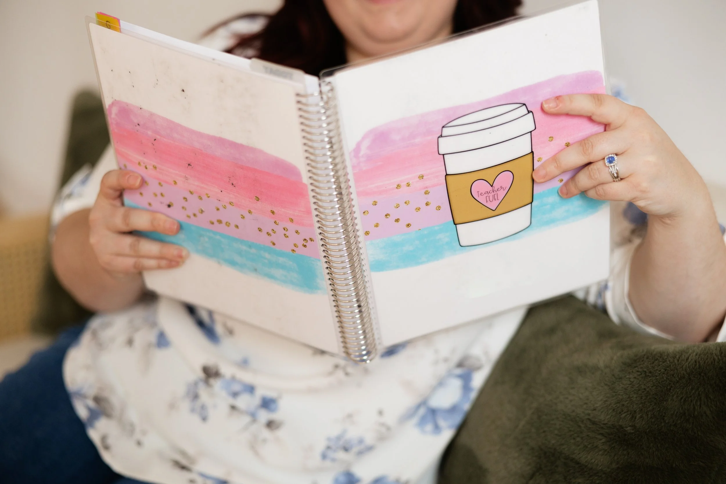 A person holding a colorful spiral-bound notebook with a pink, blue, and white abstract background and a drawing of a coffee cup with a heart and the words "Teacher Fuel" on it.