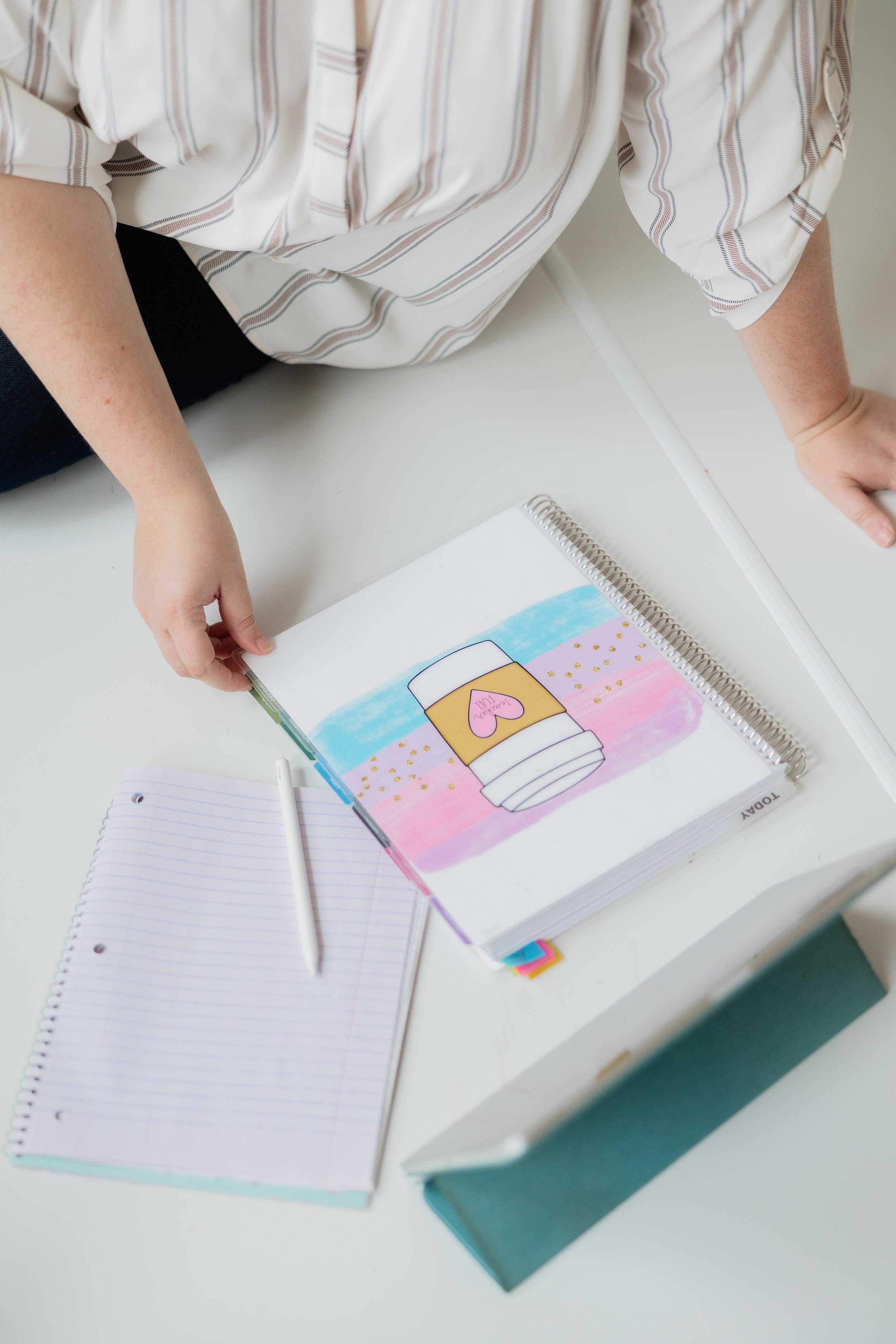 Open notebook with lined paper, white pen, closed hardcover book, and colorful illustration of a coffee cup with a pink heart.