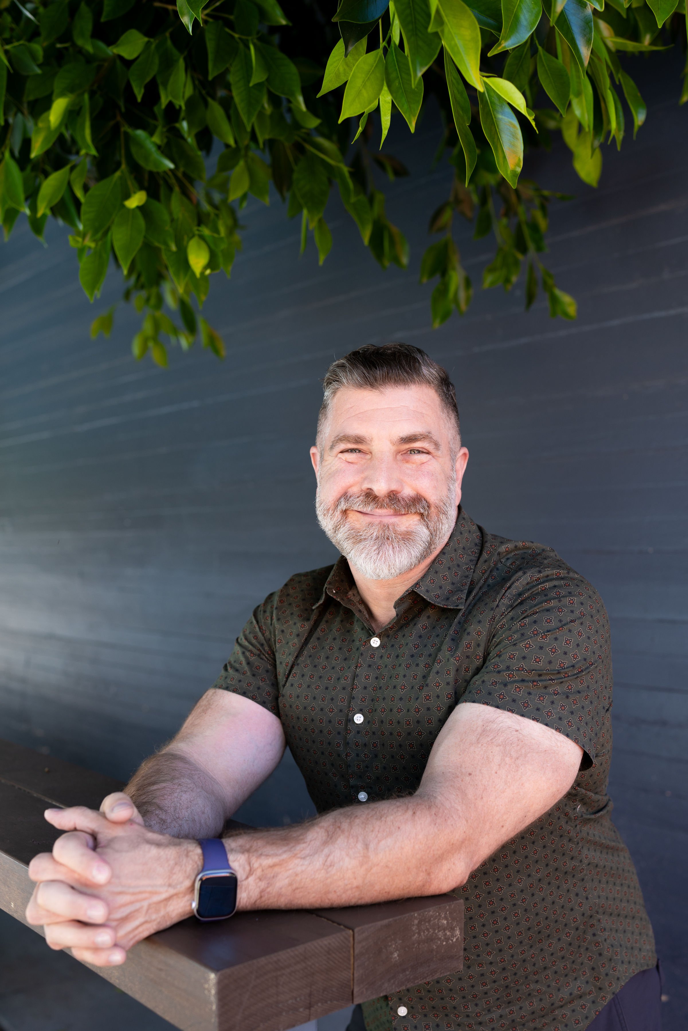 A middle-aged man with gray hair and a beard, smiling, wearing a dark shirt with a subtle pattern, sitting outdoors at a wooden table, with his hands clasped and wearing a smartwatch, in front of a background of green leaves and a dark textured wall.