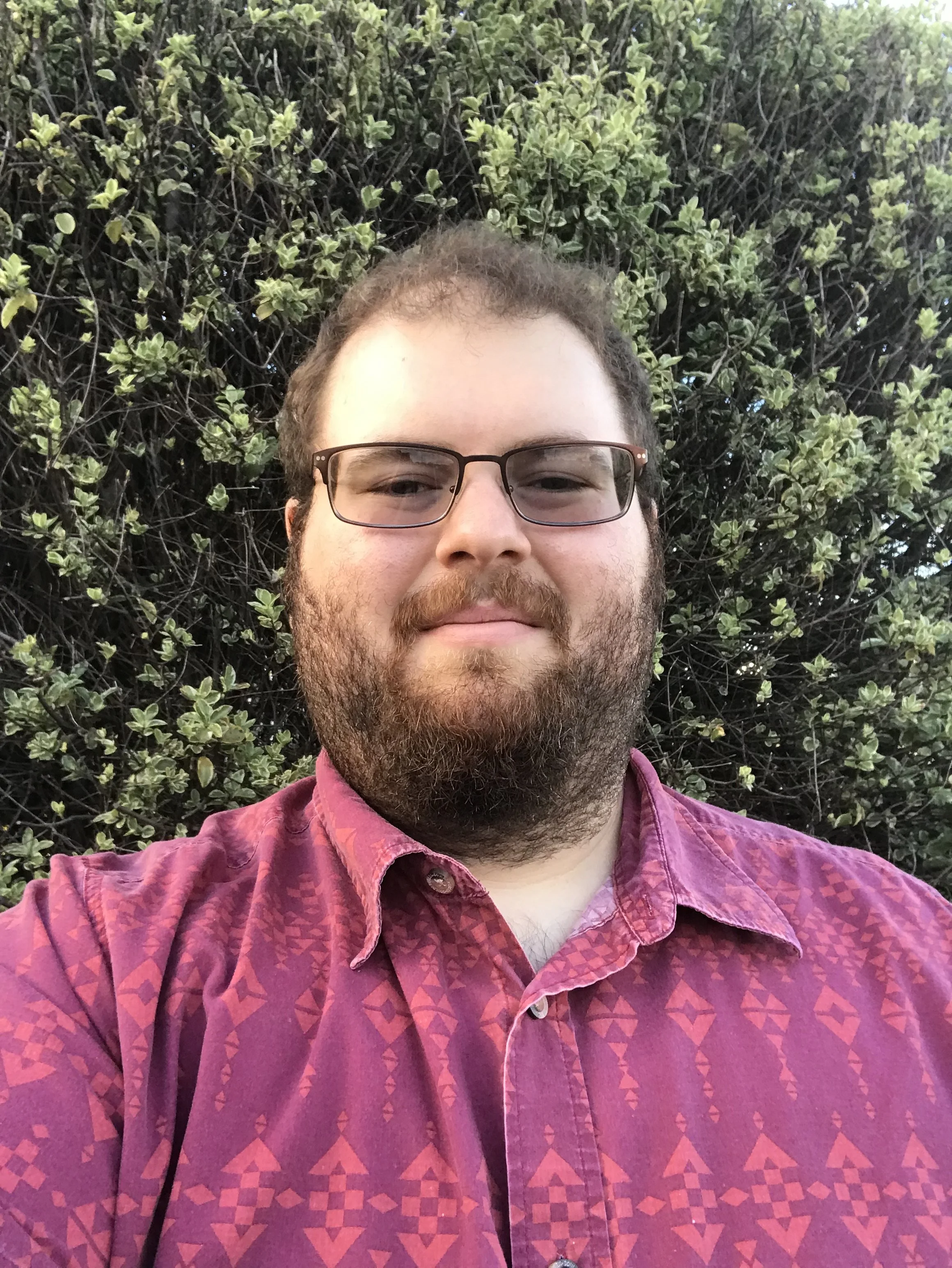 A man with glasses and a beard taking a selfie outdoors. He is wearing a pink patterned shirt. Behind him is a large bush with green leaves.  Jacob Rosen, LMFT, crisis trainings, mental health mobile crisis response, behavioral health crisis training