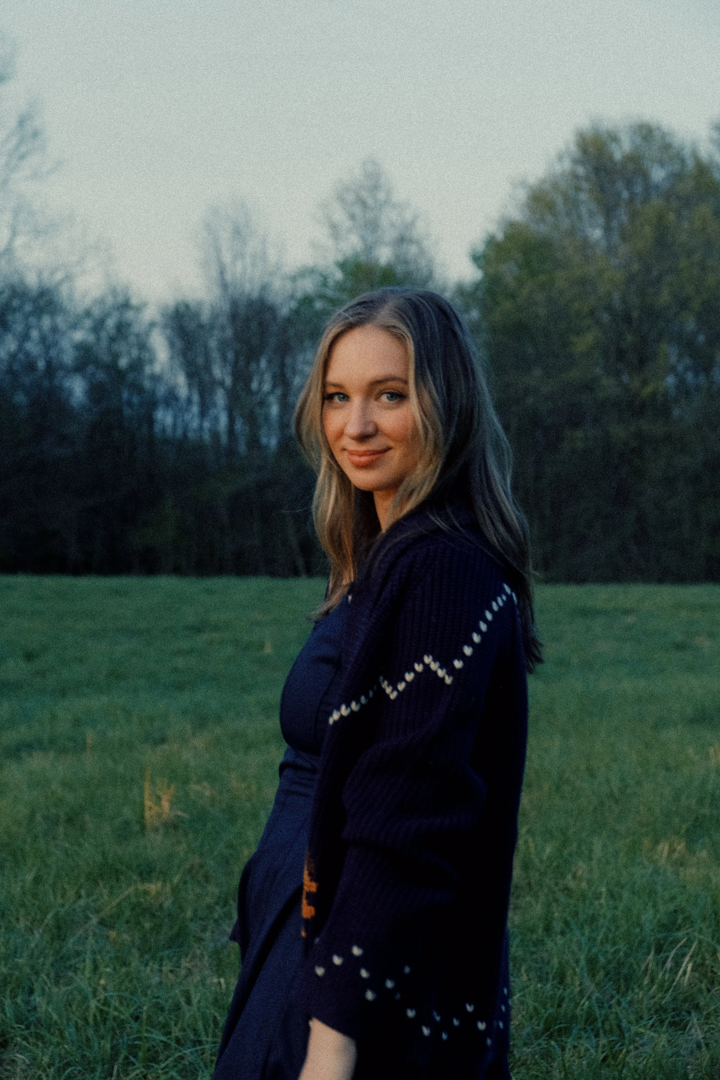 A young woman with blonde hair and blue eyes standing outdoors on a grassy field with trees in the background during dusk.
