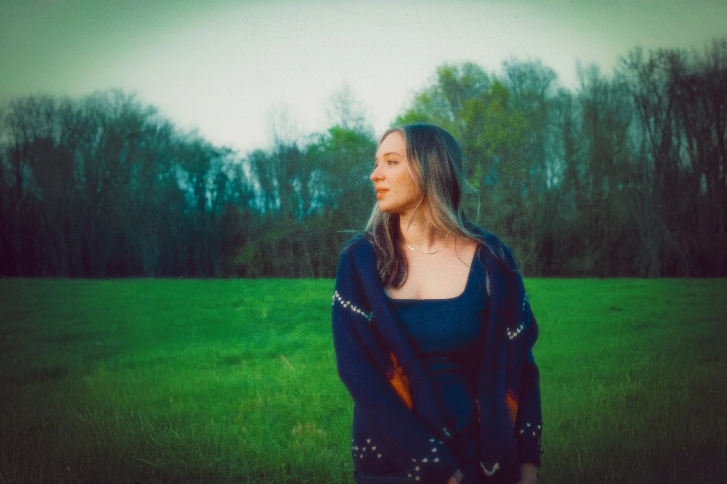 A woman with long hair wearing a dark sweater with a colorful pattern standing in a grassy field with trees in the background.